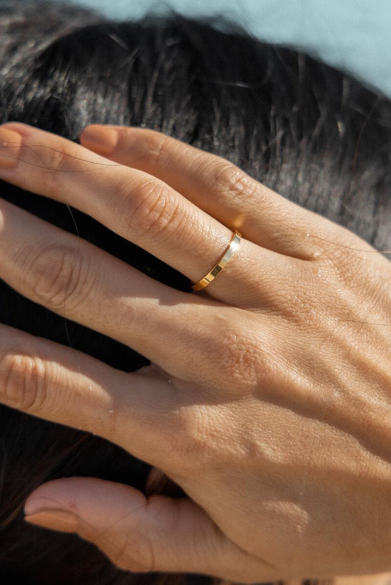 A hand wearing a simple gold-plated ring against a coastal backdrop.