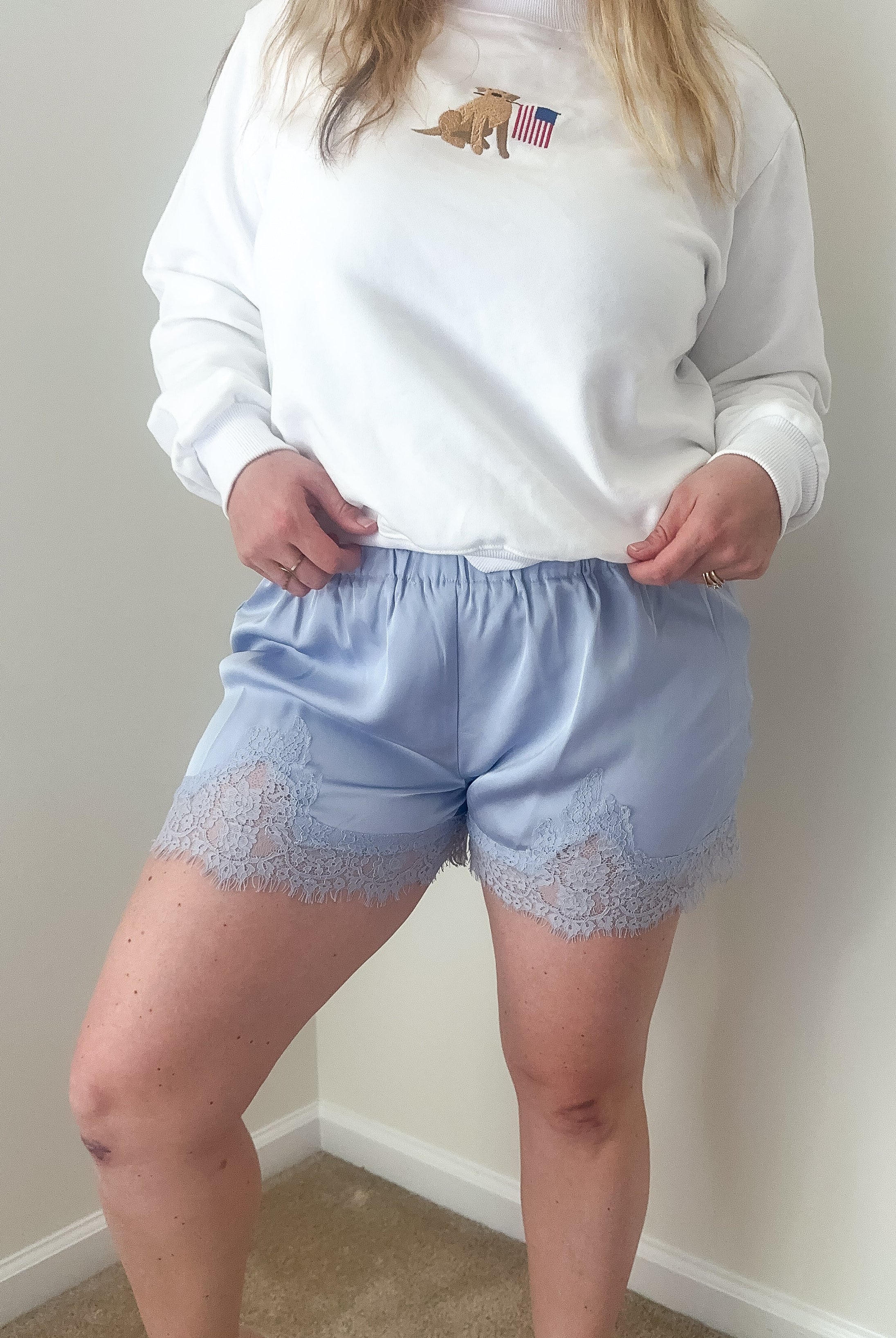 Person wearing a white sweatshirt with a logo and light blue shorts with lace trim against a plain wall.