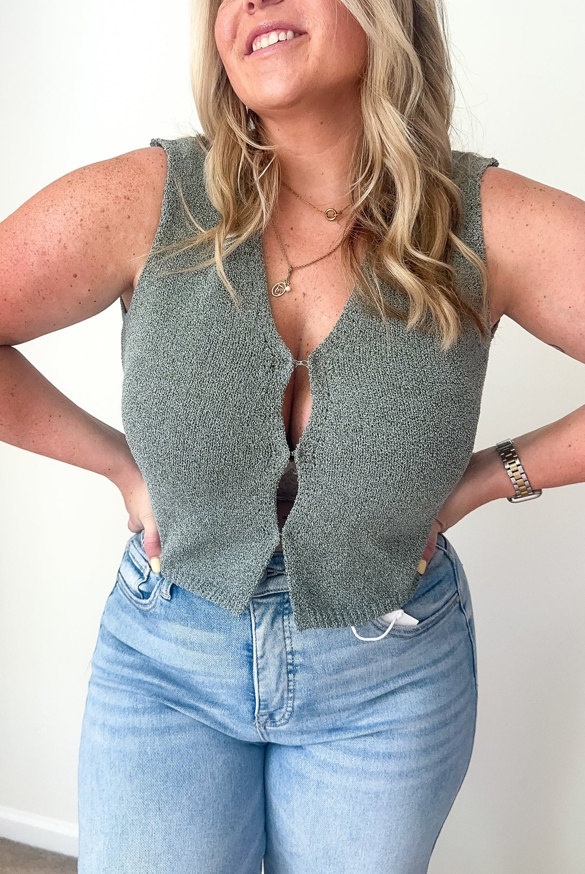 A sleeveless olive green knit vest cardigan with a deep V-neckline and hook-and-eye closure.