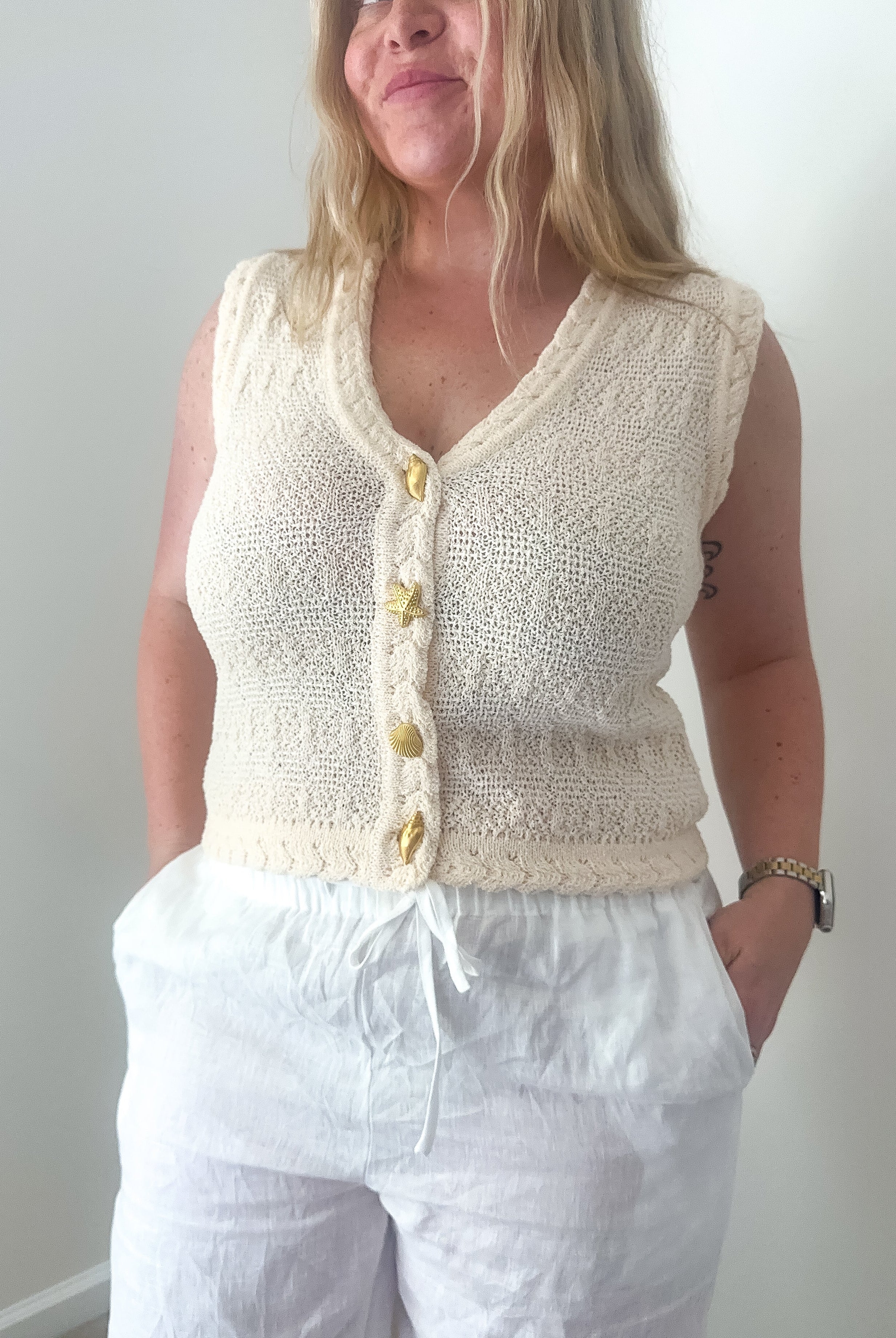 Woman wearing a cream knitted vest with buttons against a plain background