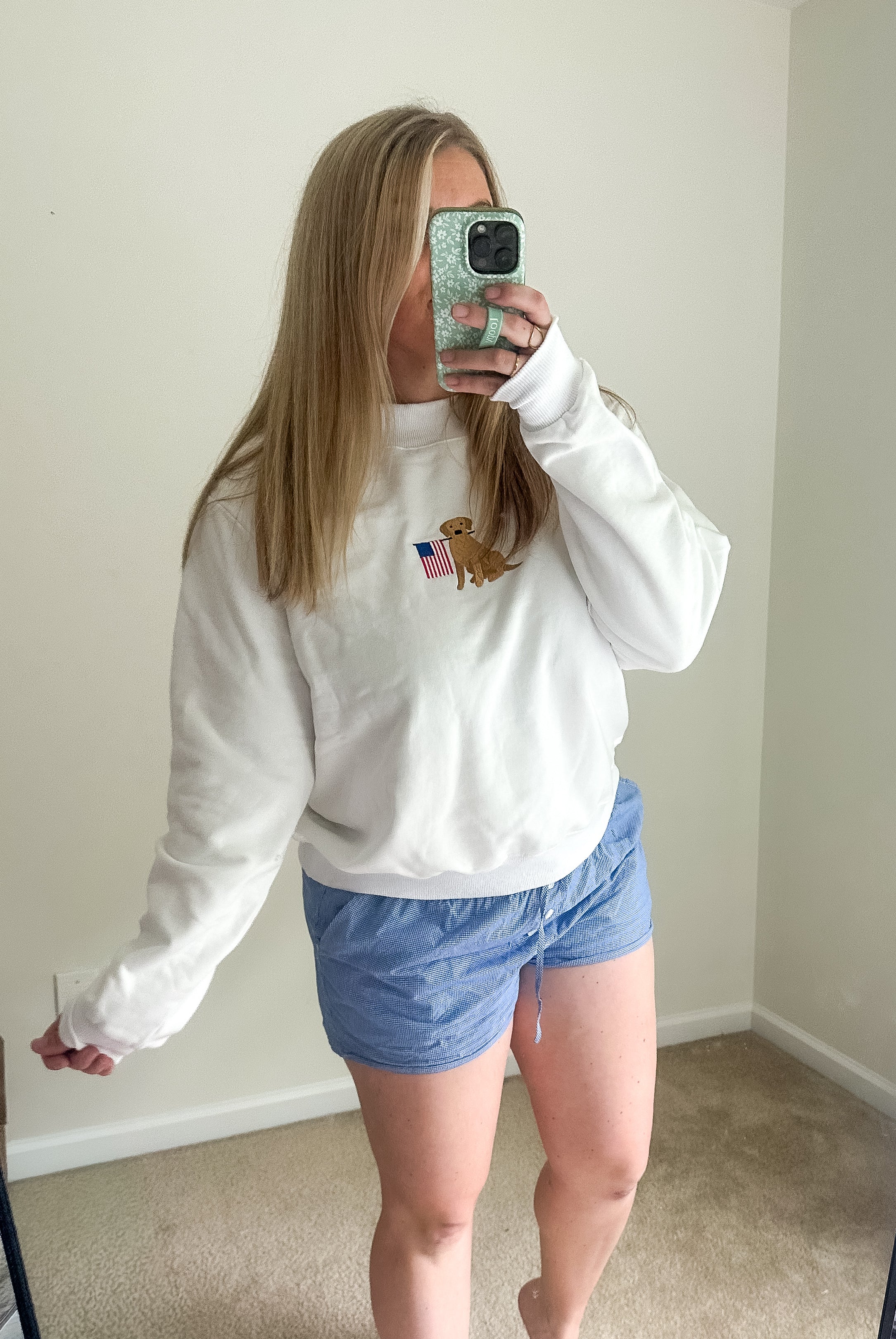 Person wearing a white sweatshirt with a logo and blue shorts taking a mirror selfie.