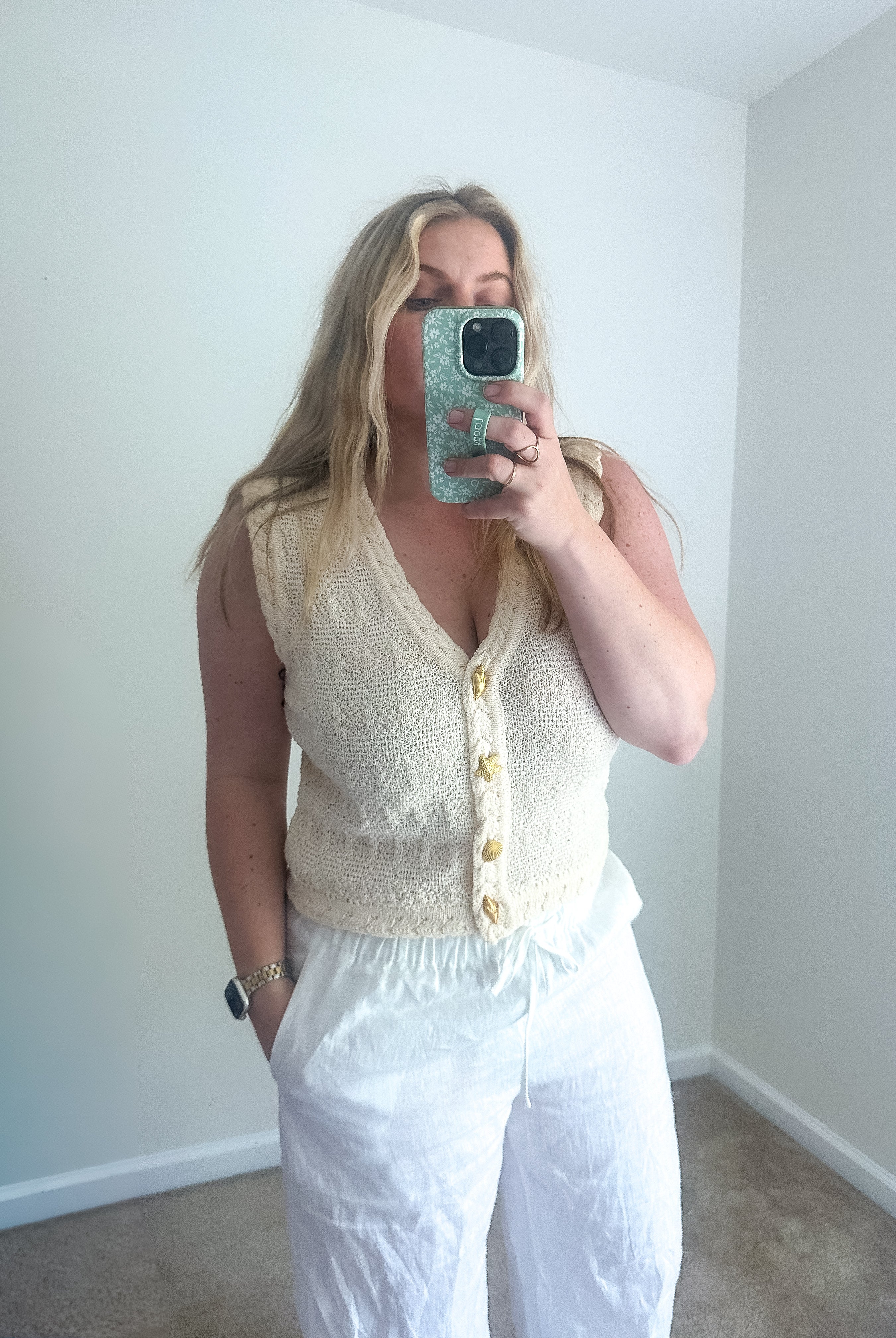 Woman taking a mirror selfie wearing a white sleeveless top and pants in a room with white walls.