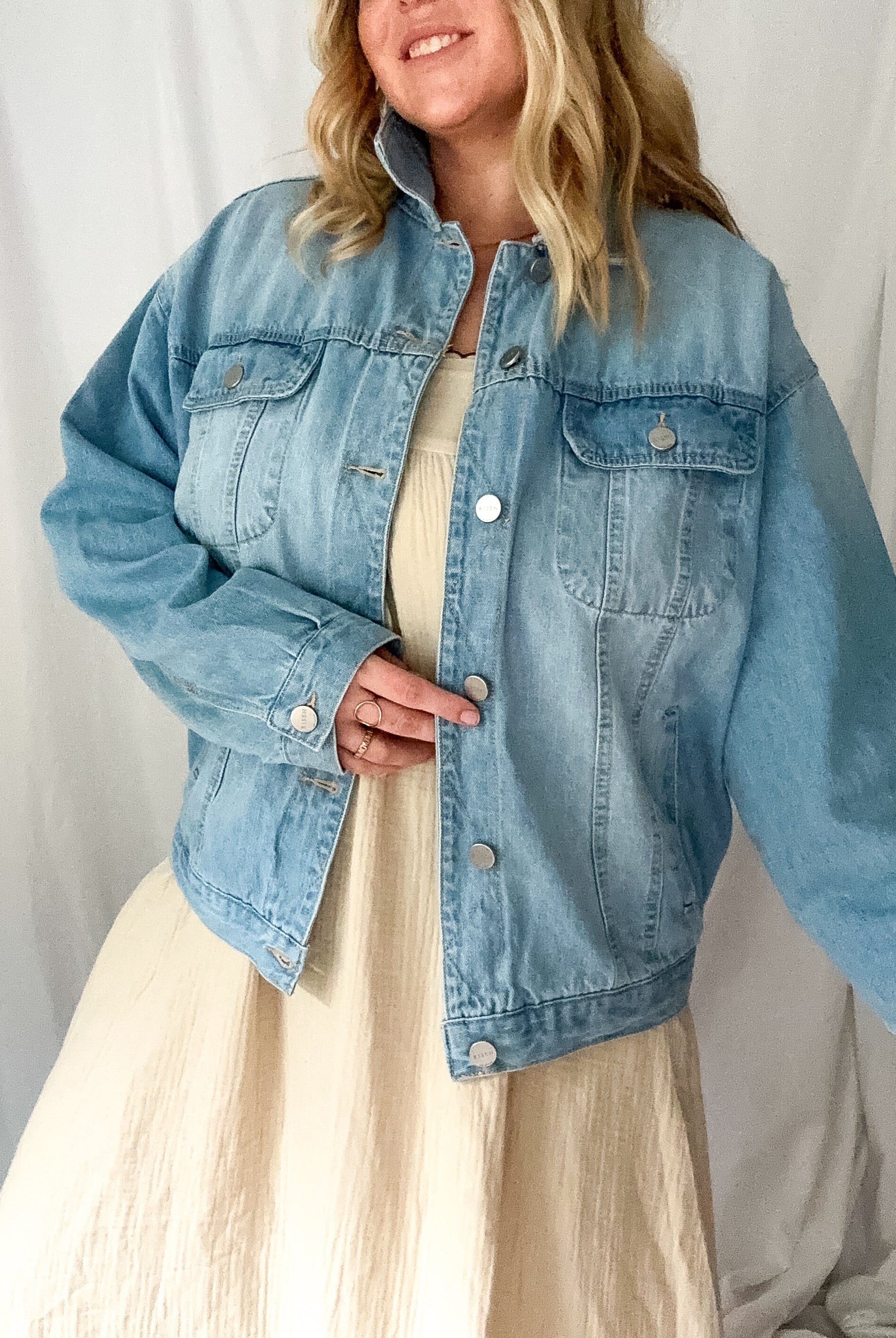 A medium wash denim jacket with an oversized fit, worn open over a cream dress.
