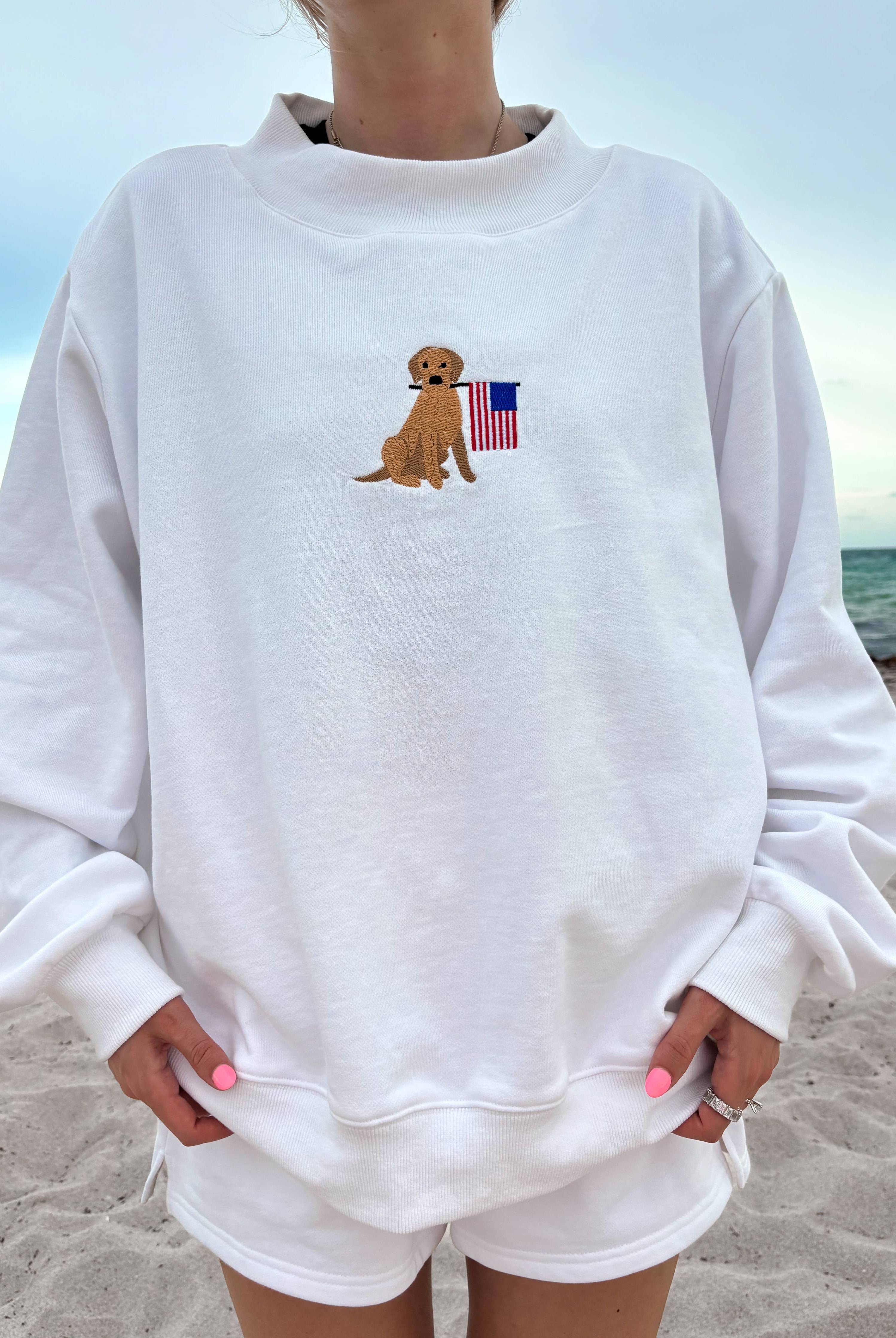 Person wearing a white sweatshirt with a dog and American flag design on a beach.