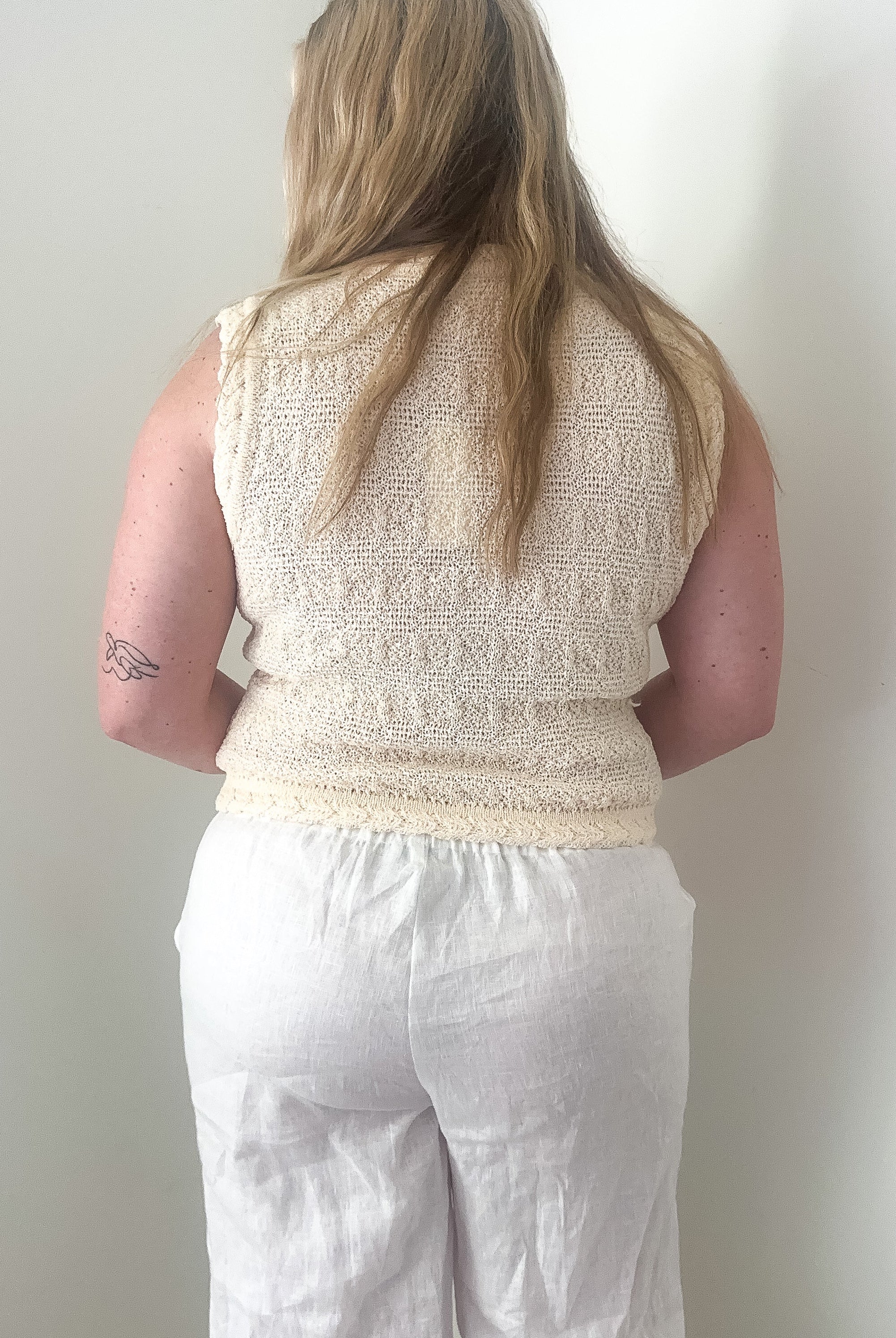 Person wearing a beige sleeveless top and white pants against a plain wall.