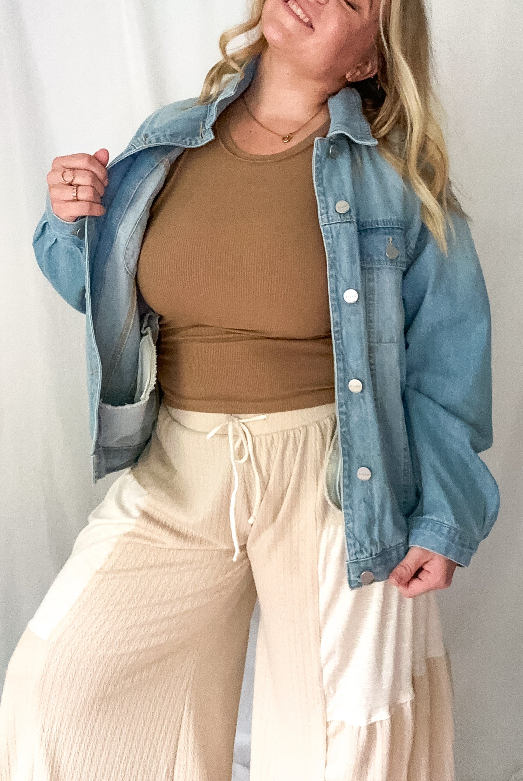 Woman wearing a denim jacket, brown top, and beige pants against a white background