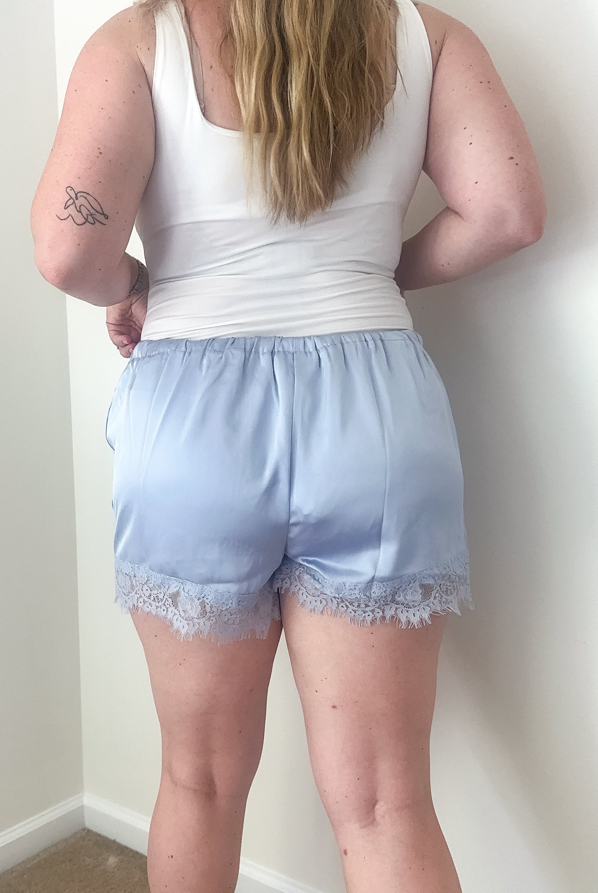 Person wearing a white tank top and light blue shorts with lace trim against a plain wall.