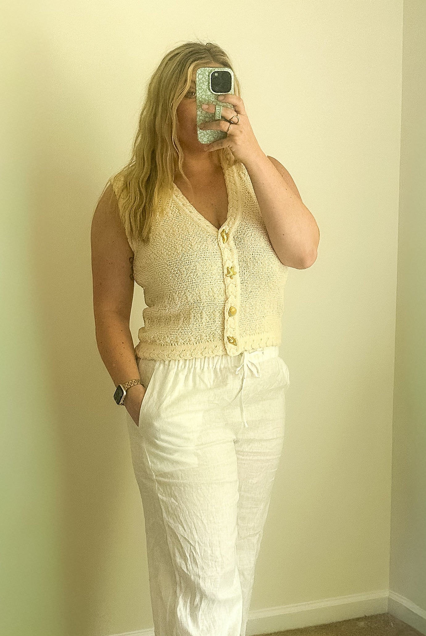 Person taking a mirror selfie wearing a yellow sleeveless top and white pants in a room with light green walls.