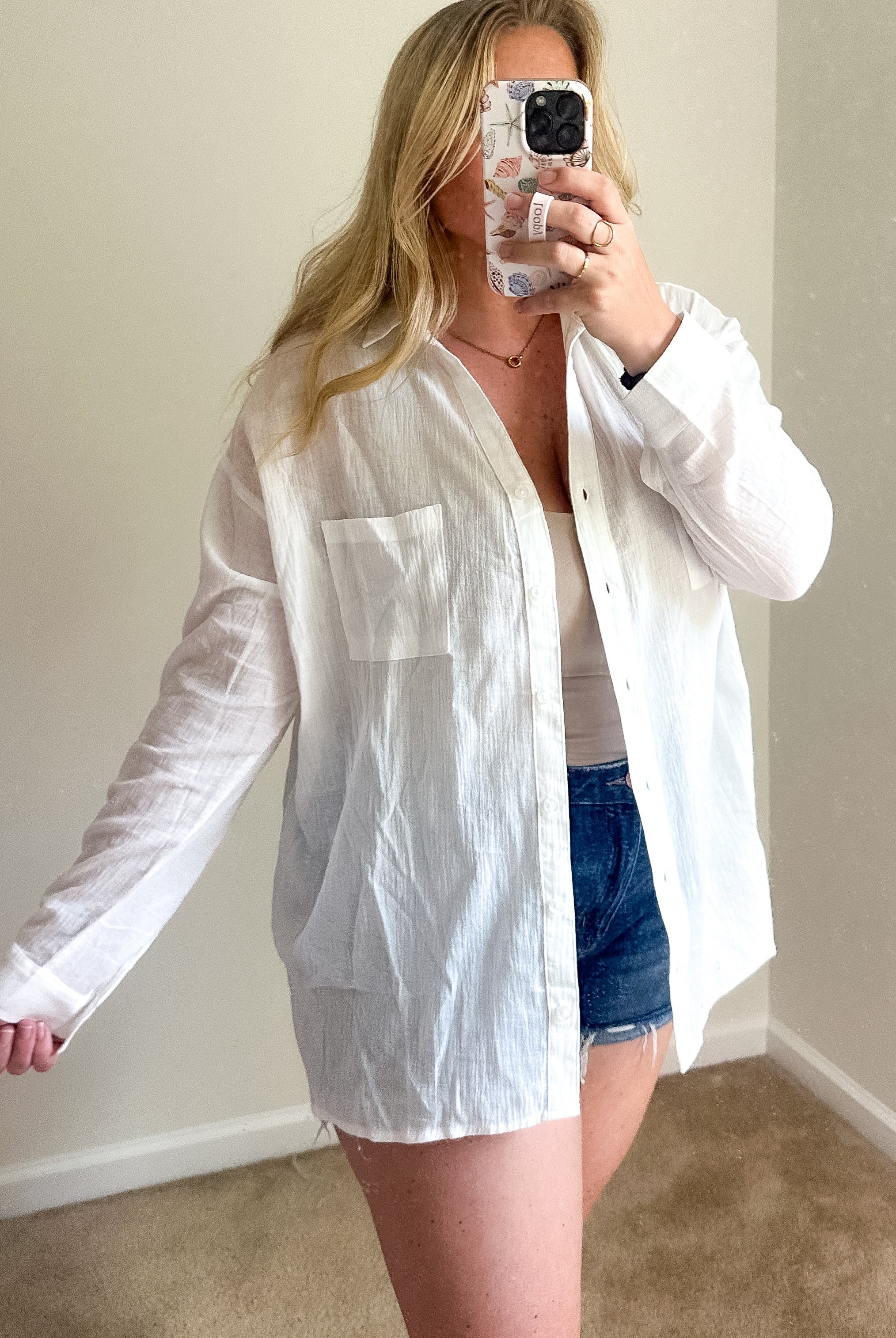 A woman takes a mirror selfie wearing an oversized white long-sleeve button-up shirt over a white top and denim shorts in a room with beige walls and carpet.