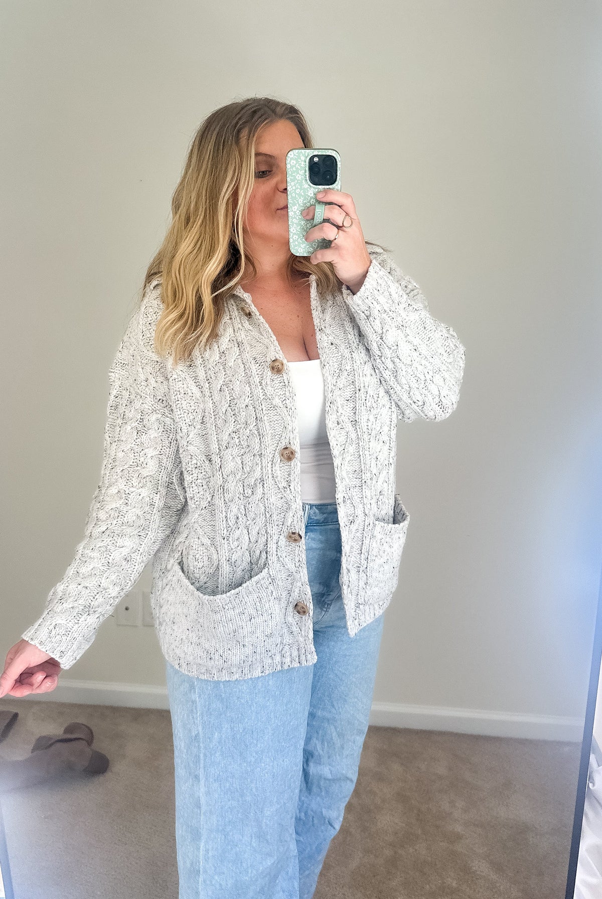 Woman taking a mirror selfie wearing a patterned cardigan and light blue jeans.