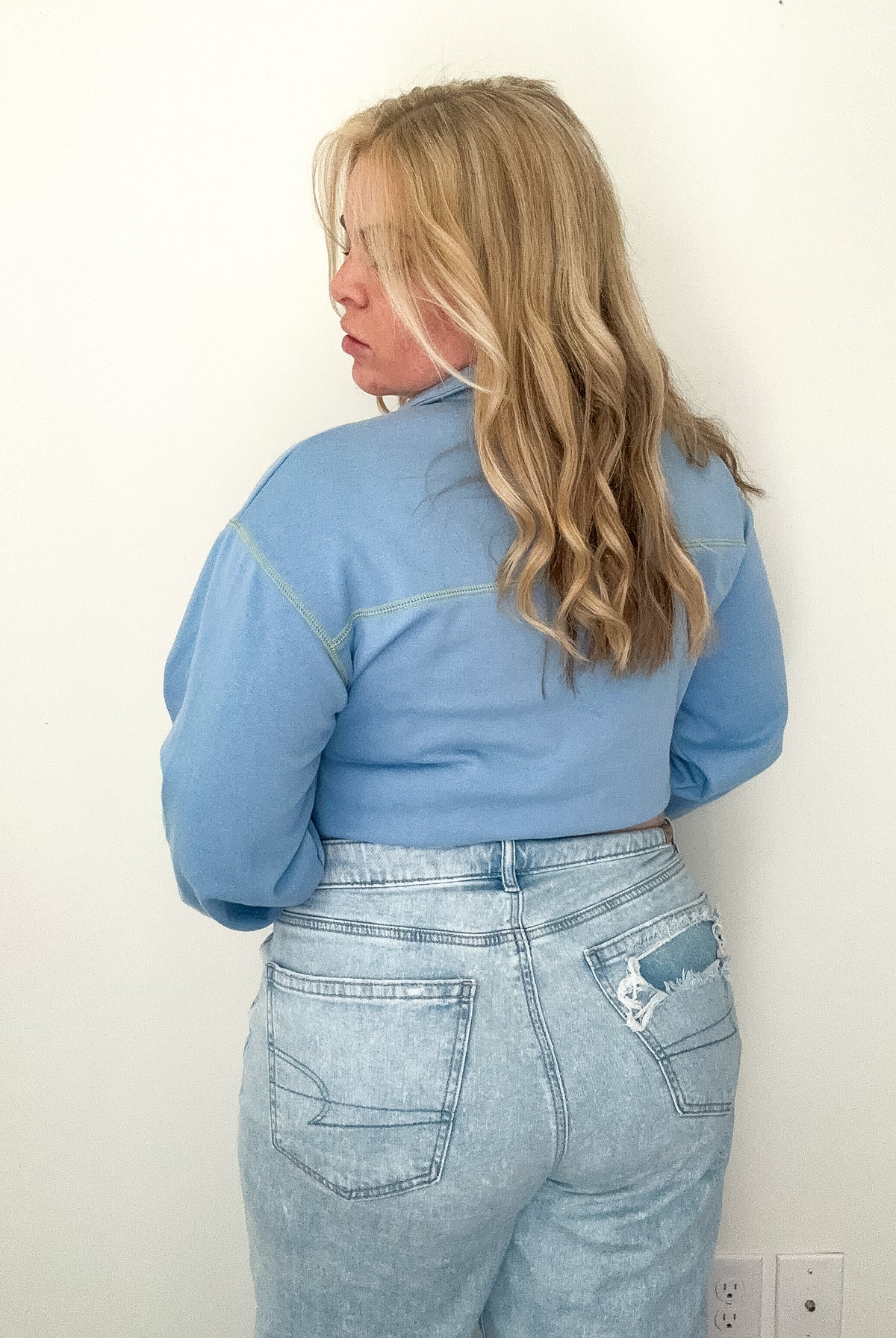 Person wearing a blue jacket and jeans against a white wall.