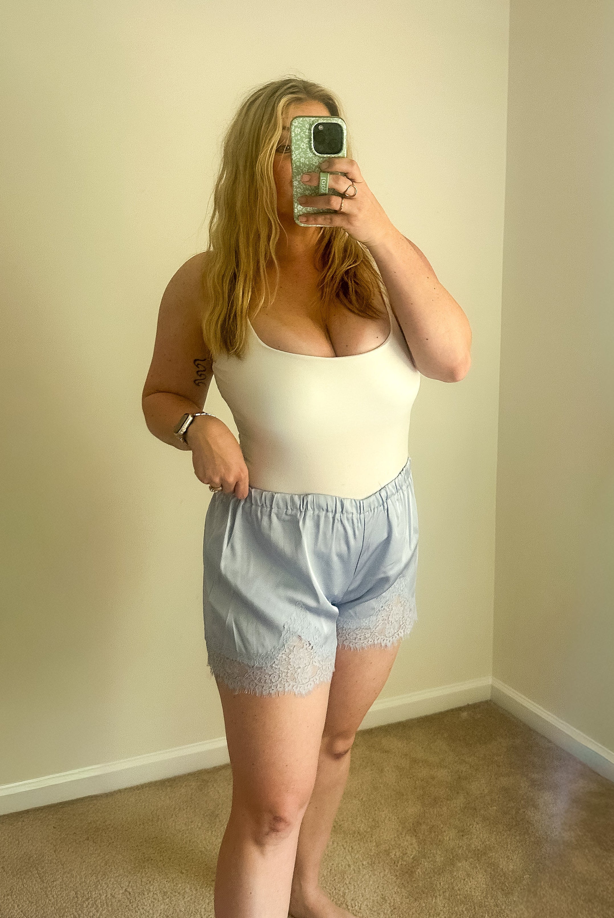 Person taking a mirror selfie wearing a white tank top and light blue shorts in a room with beige walls and carpet.