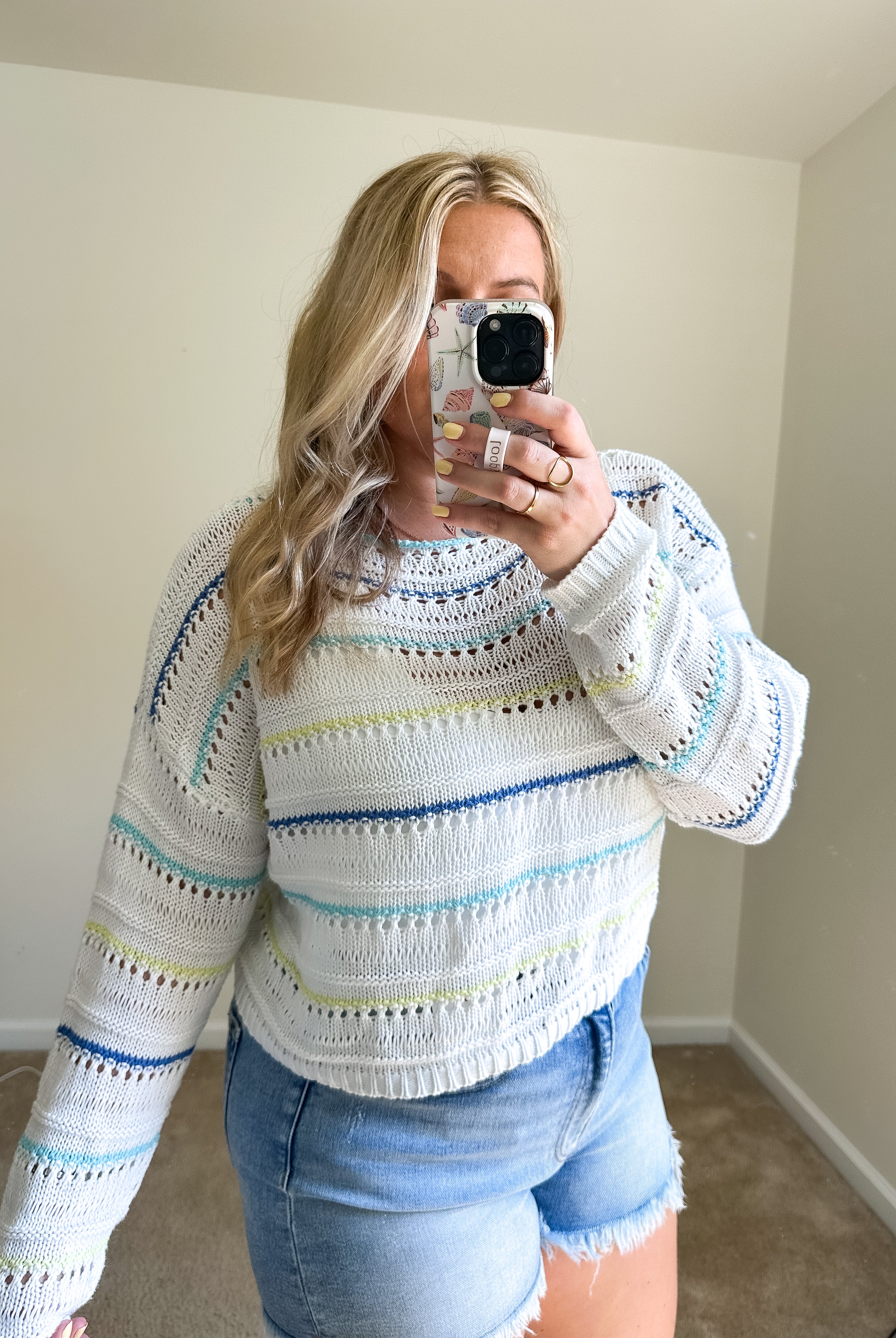 A woman takes a mirror selfie wearing a white knit sweater with horizontal stripes in light blue, yellow, and green, paired with denim shorts in a neutral indoor room.