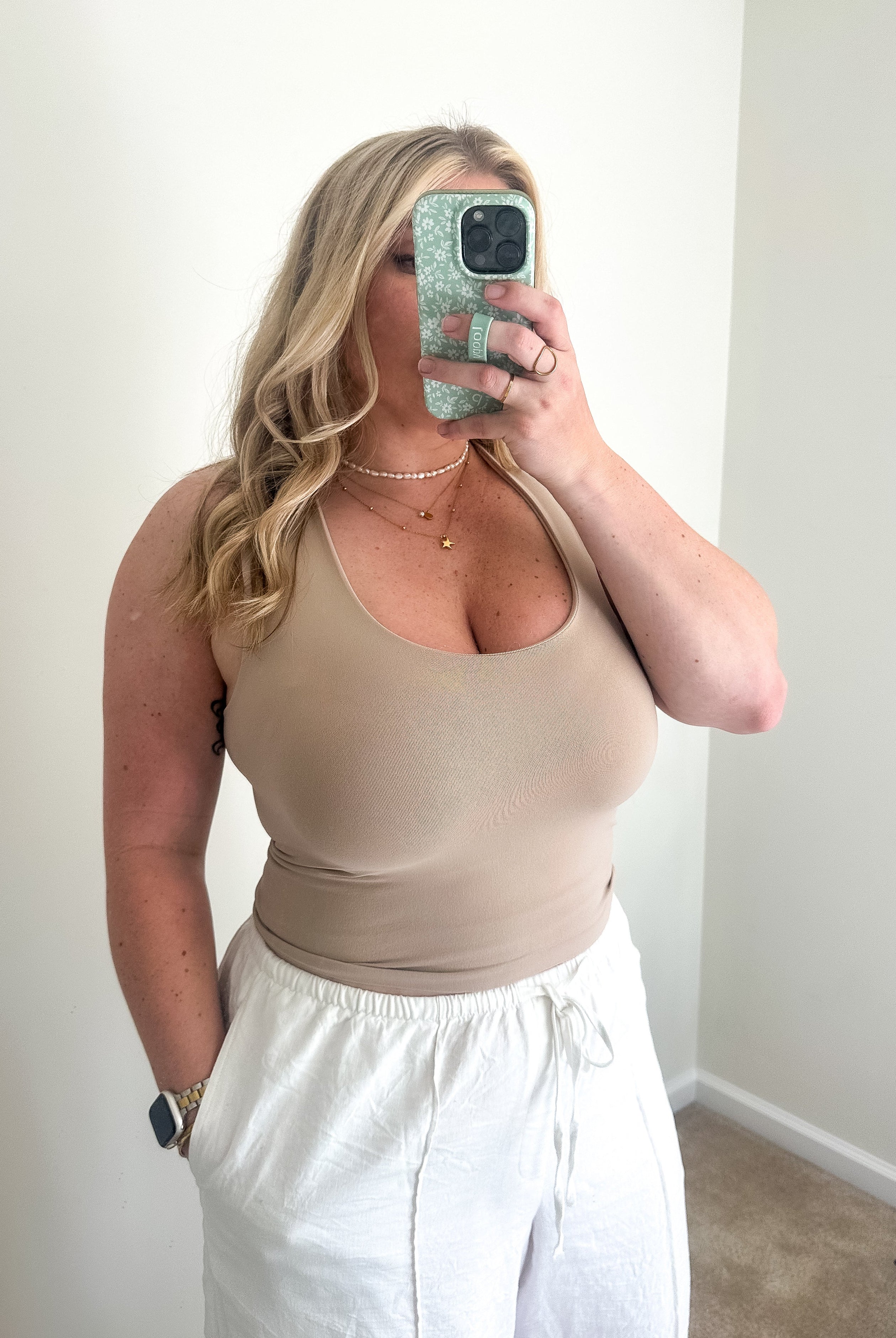 Woman taking a mirror selfie wearing a beige tank top and white pants.
