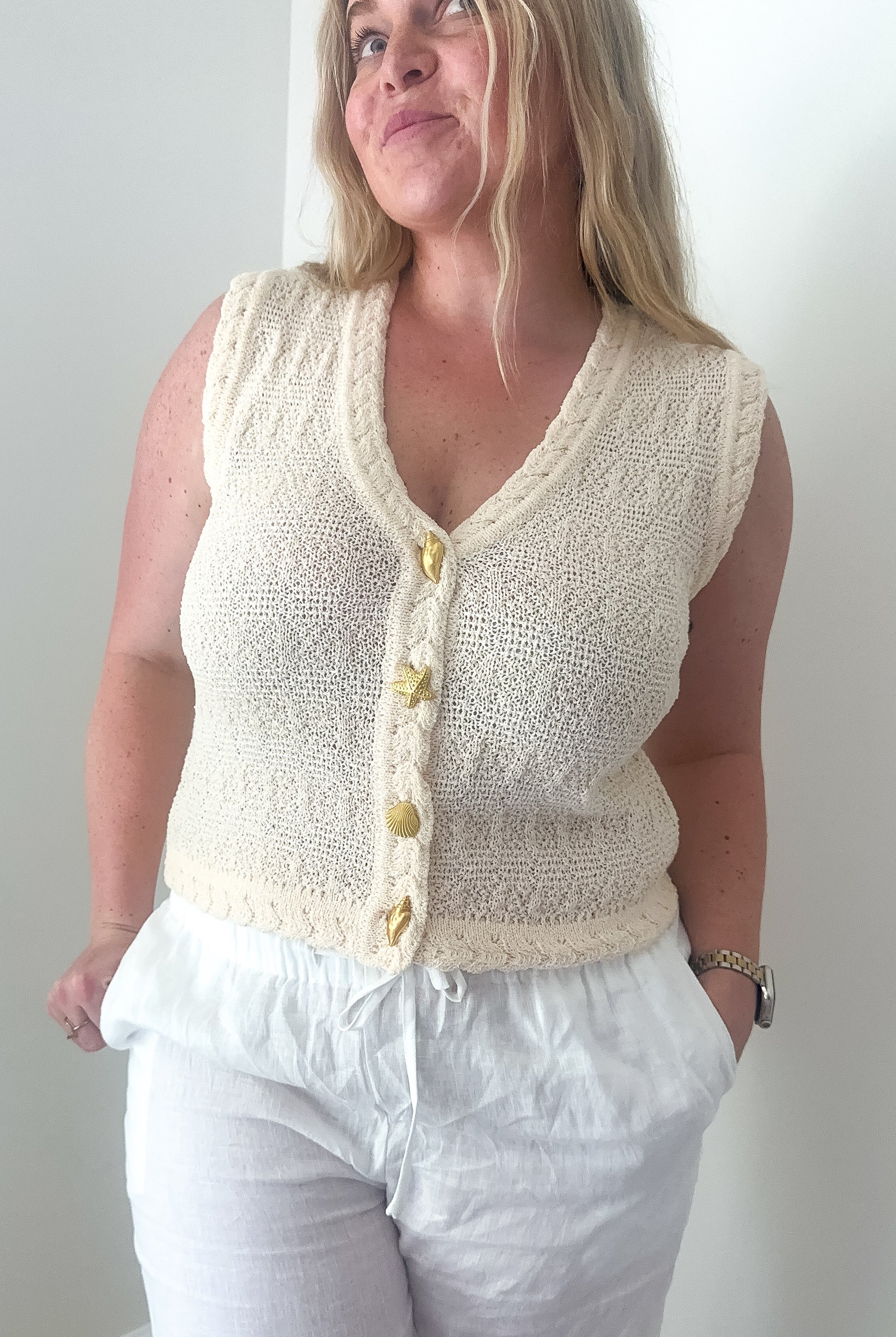 Woman wearing a cream knitted vest over a white top and pants against a plain background