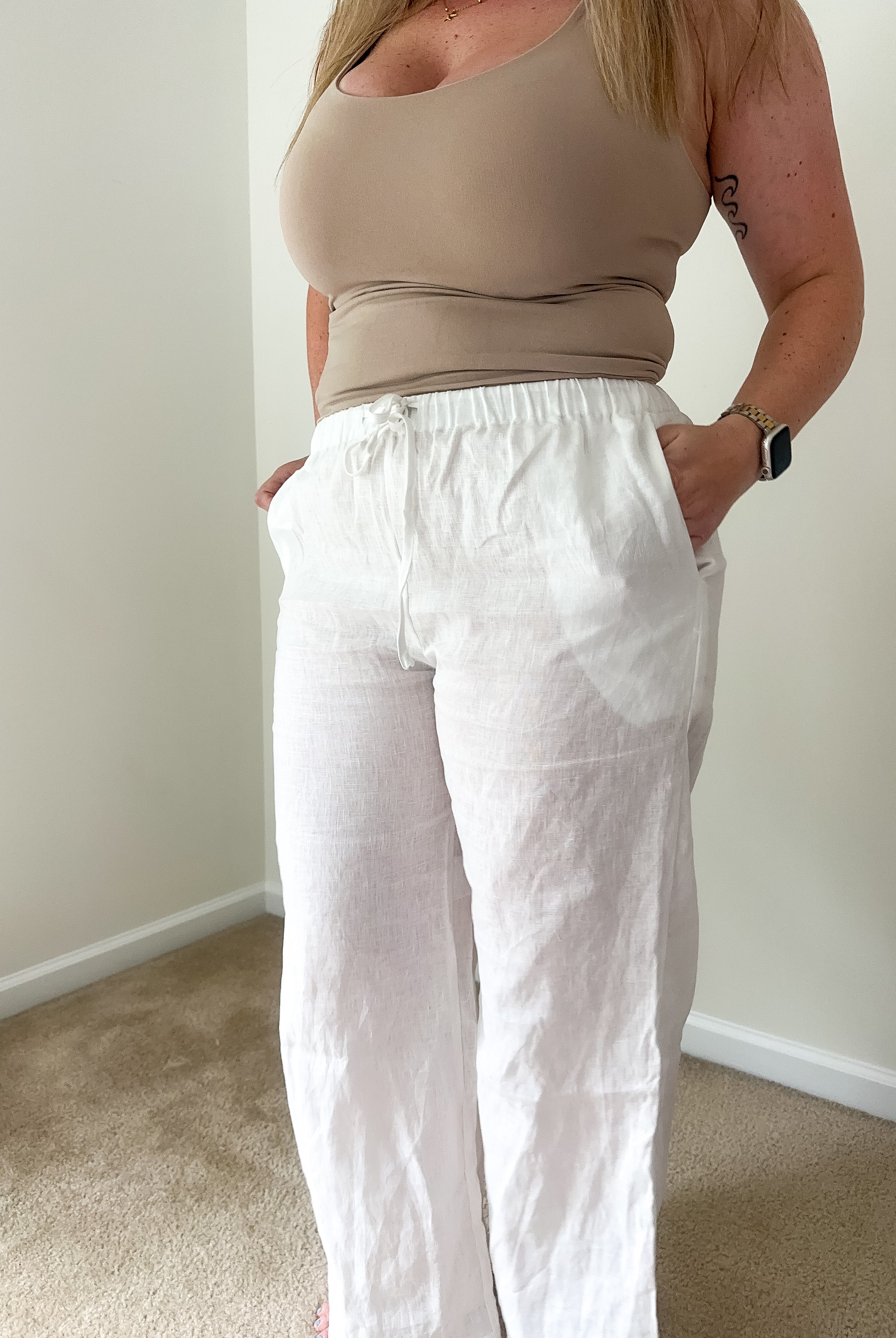 Person wearing a beige top and white pants against a plain wall.