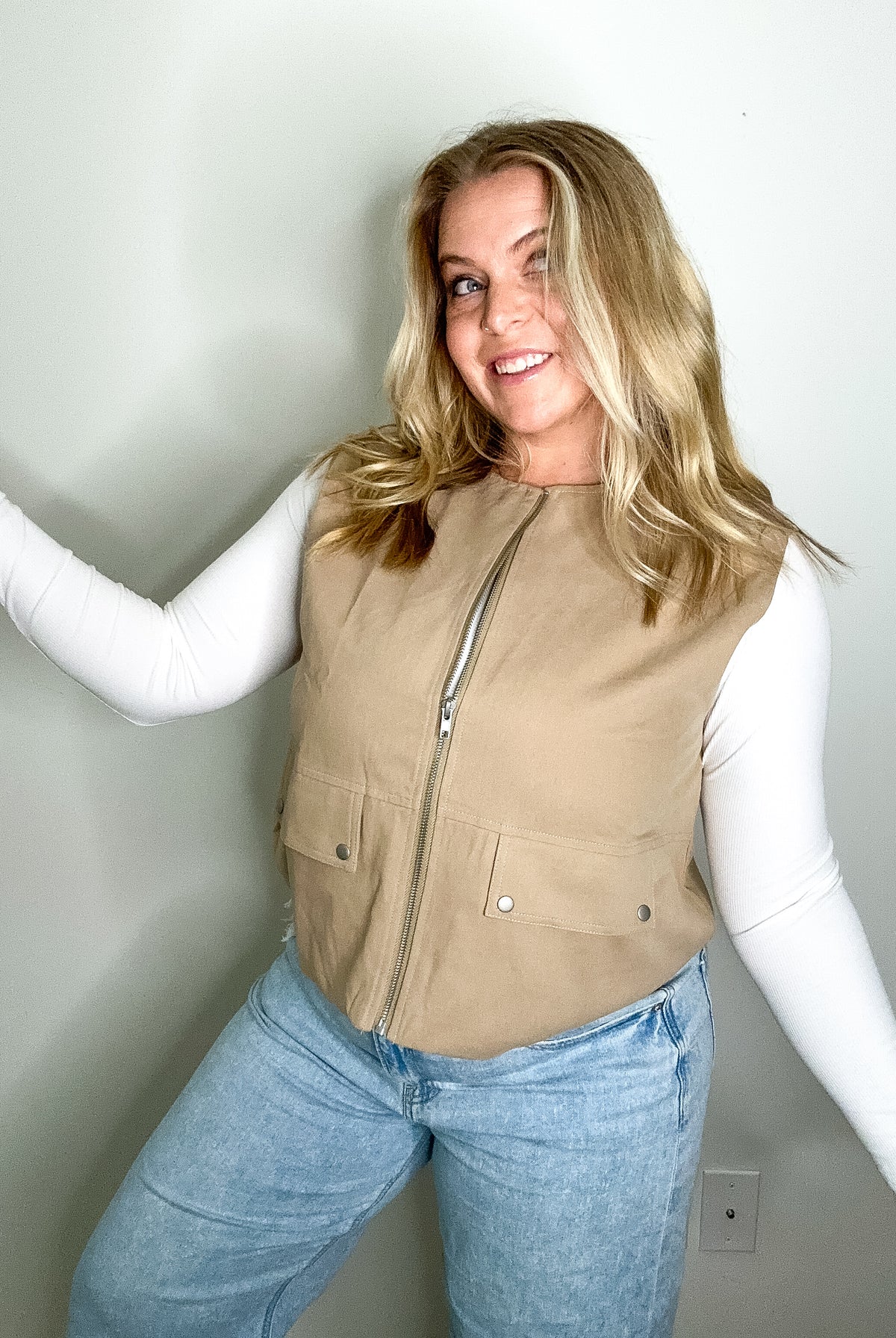Woman wearing a beige vest over a white long-sleeve shirt and blue jeans against a plain background