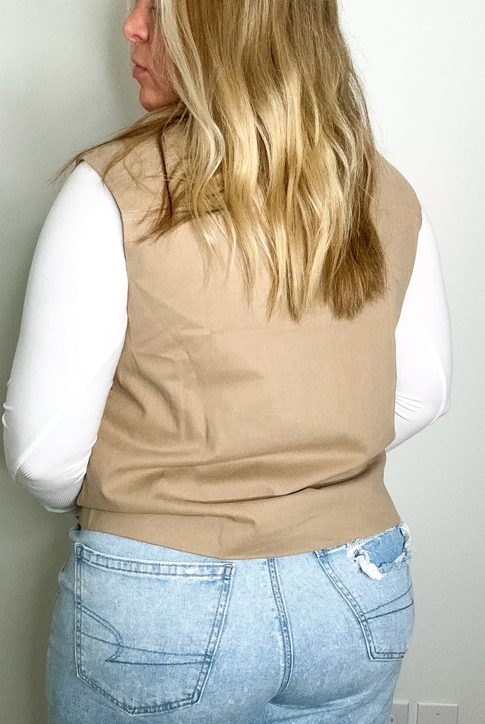 Person wearing a beige vest over a white shirt and blue jeans against a plain wall.