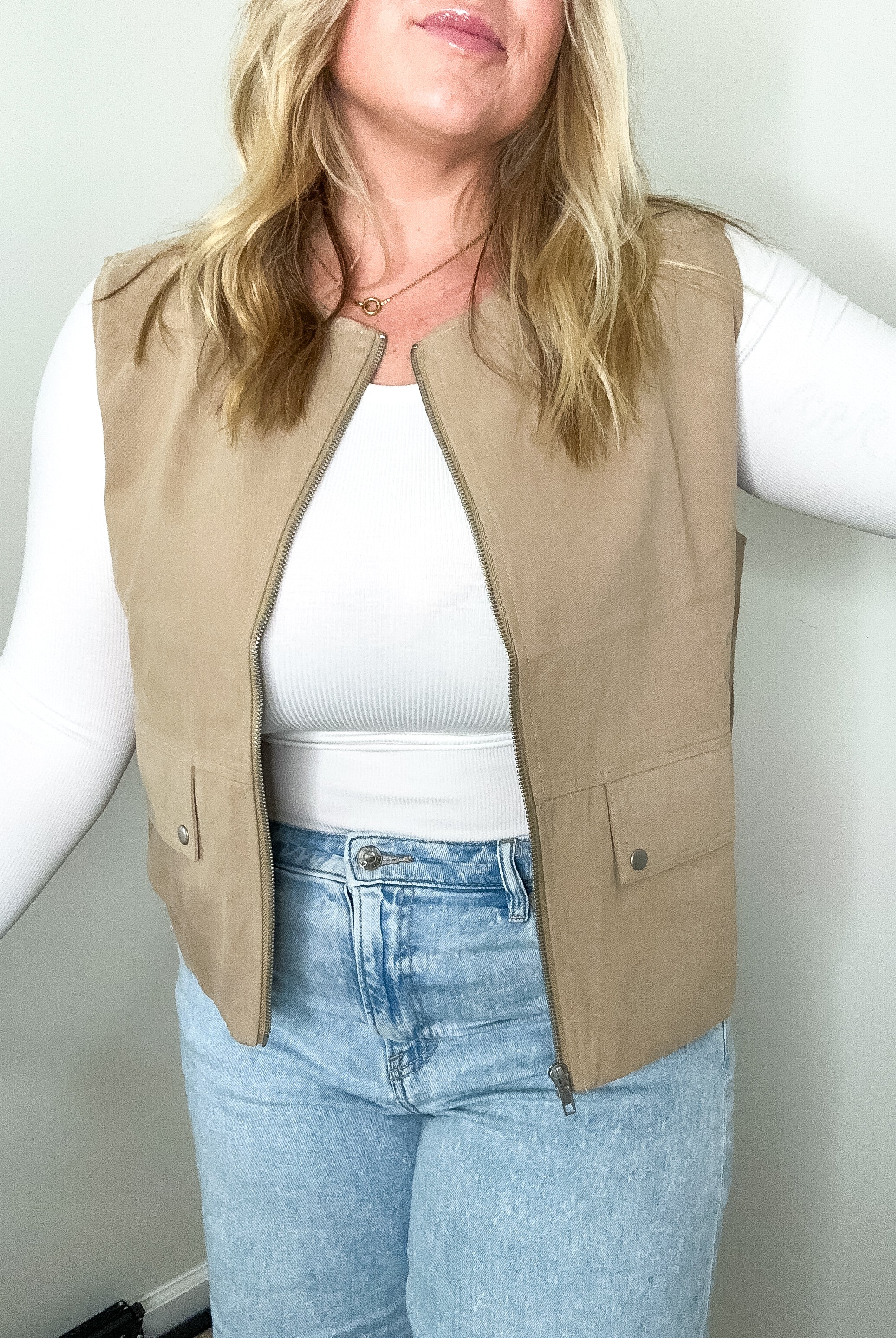 Person wearing a beige vest over a white shirt and blue jeans against a plain background
