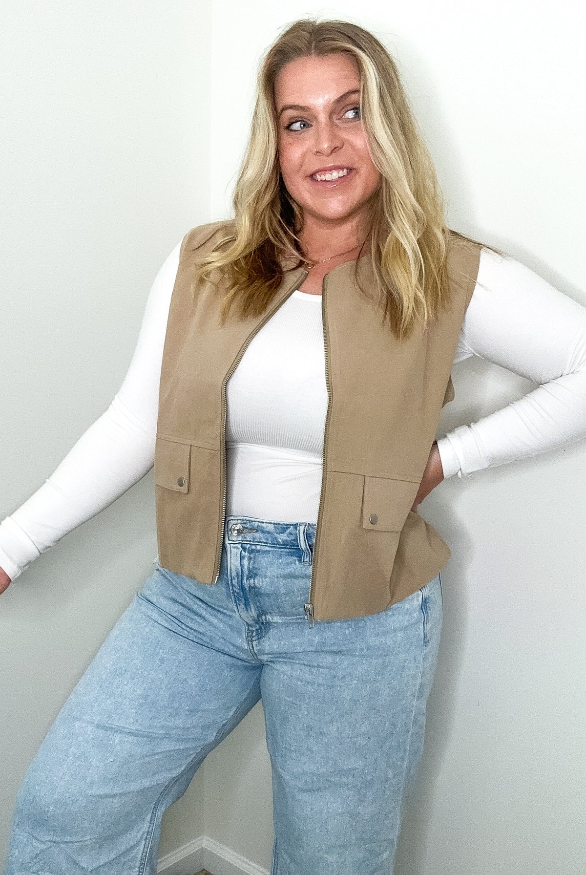 Woman wearing a beige vest, white long-sleeve shirt, and blue jeans against a plain background