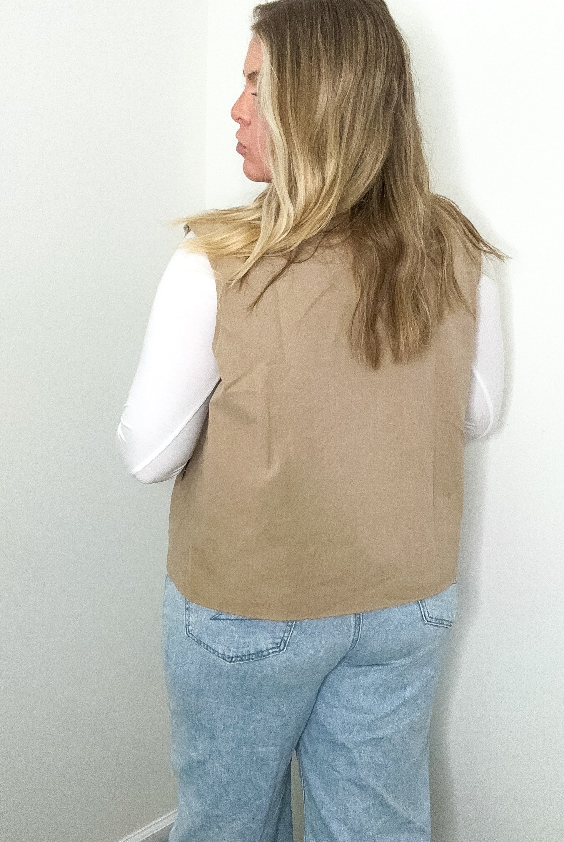 Person wearing a beige vest and light blue jeans against a white wall