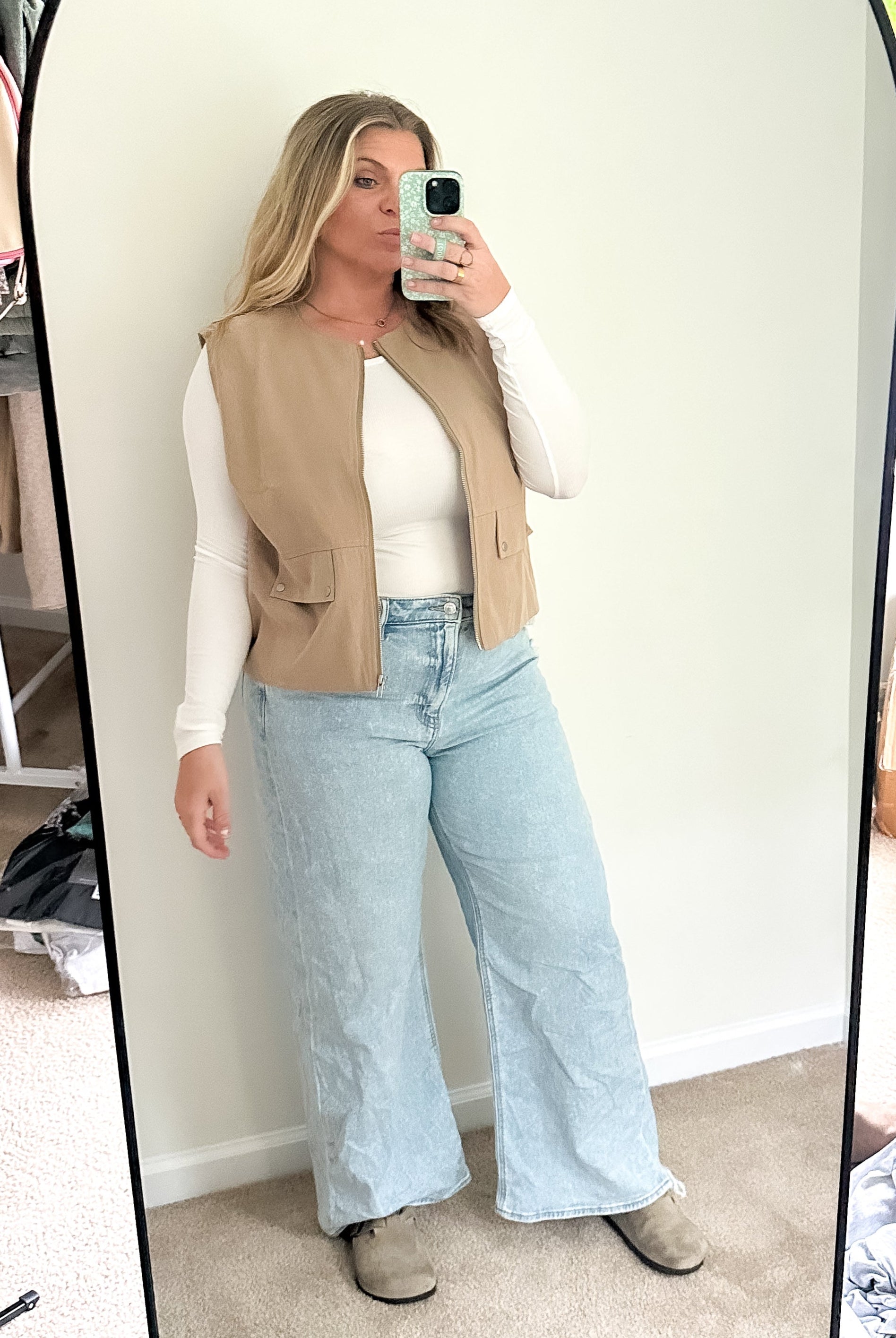 Person taking a mirror selfie wearing a beige vest, white shirt, and light blue jeans in a room with a window and boxes.