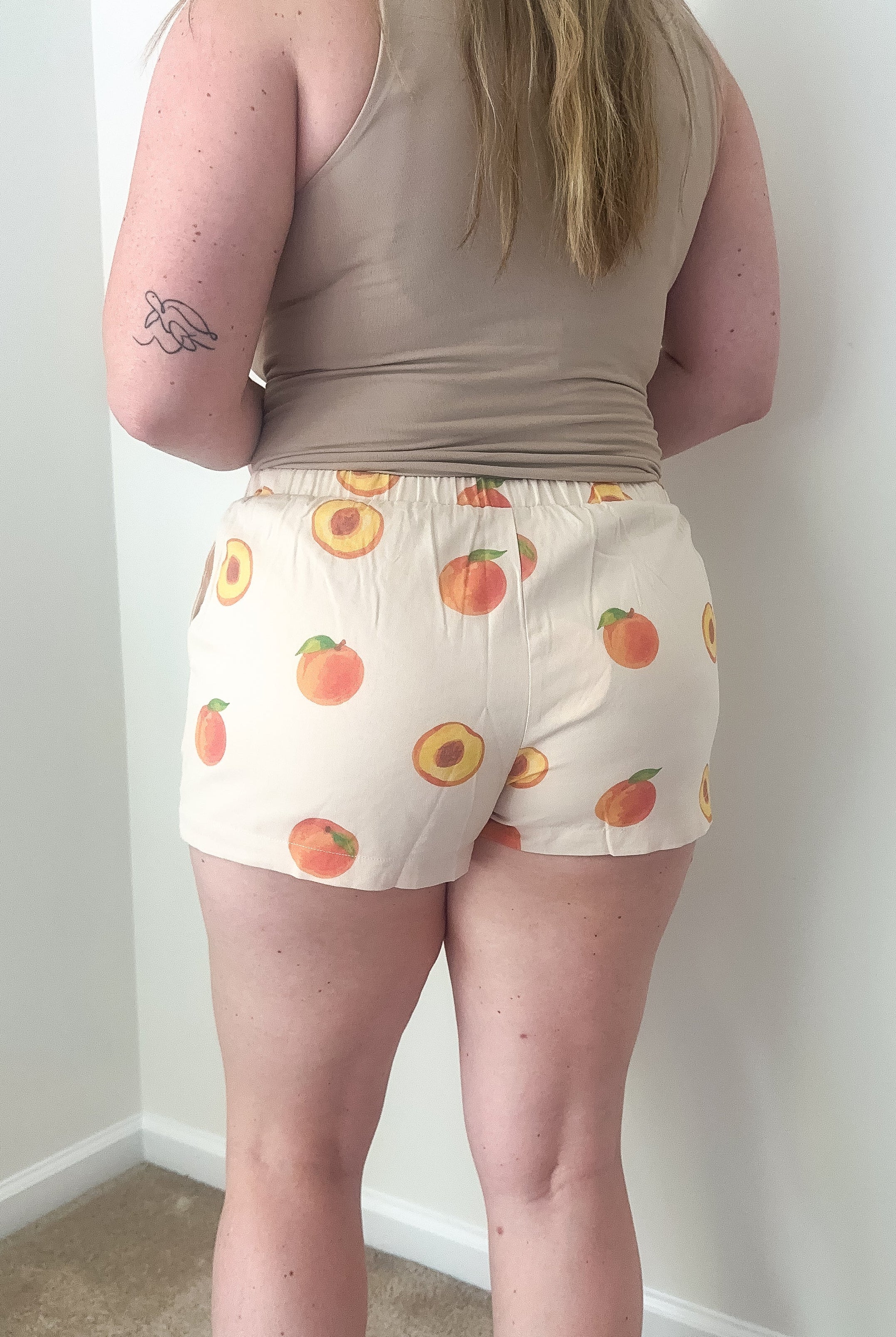 Person wearing a beige tank top and shorts with peach pattern against a white wall.