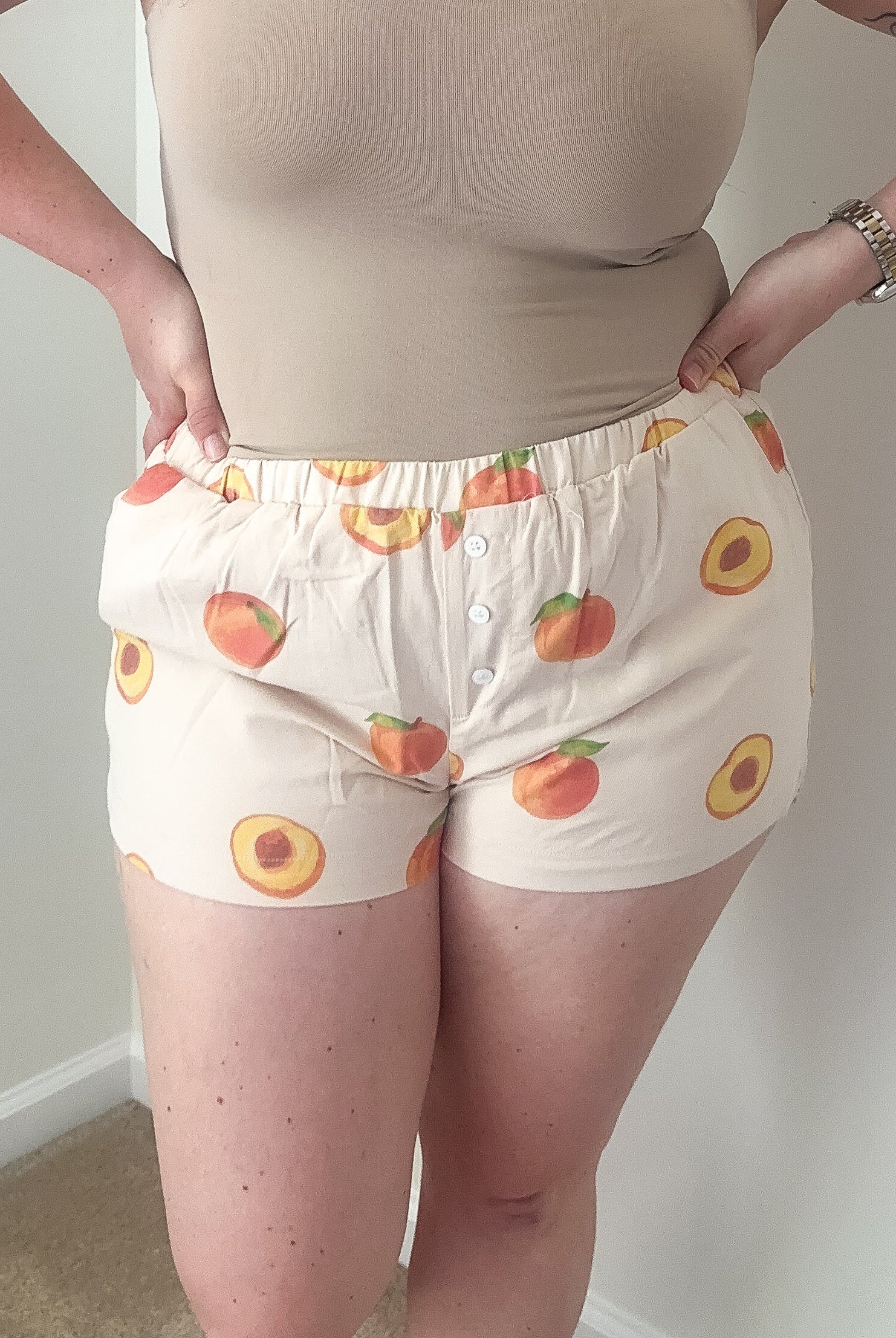 Woman wearing a beige tank top and patterned shorts against a plain wall.