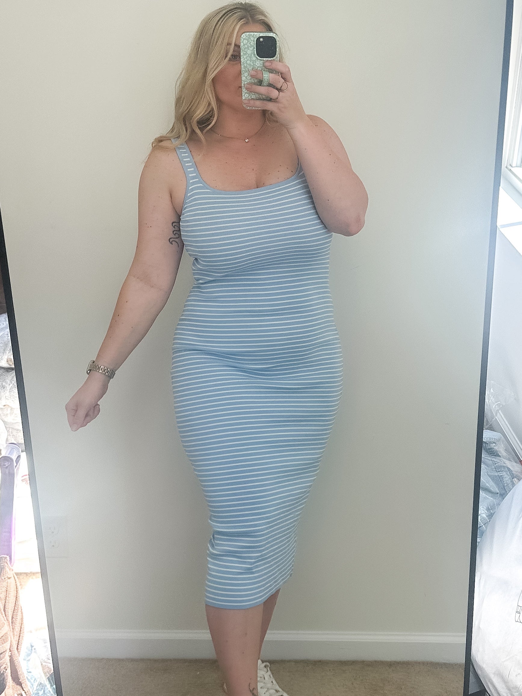 Person wearing a blue striped dress taking a mirror selfie in a room with a window and furniture.