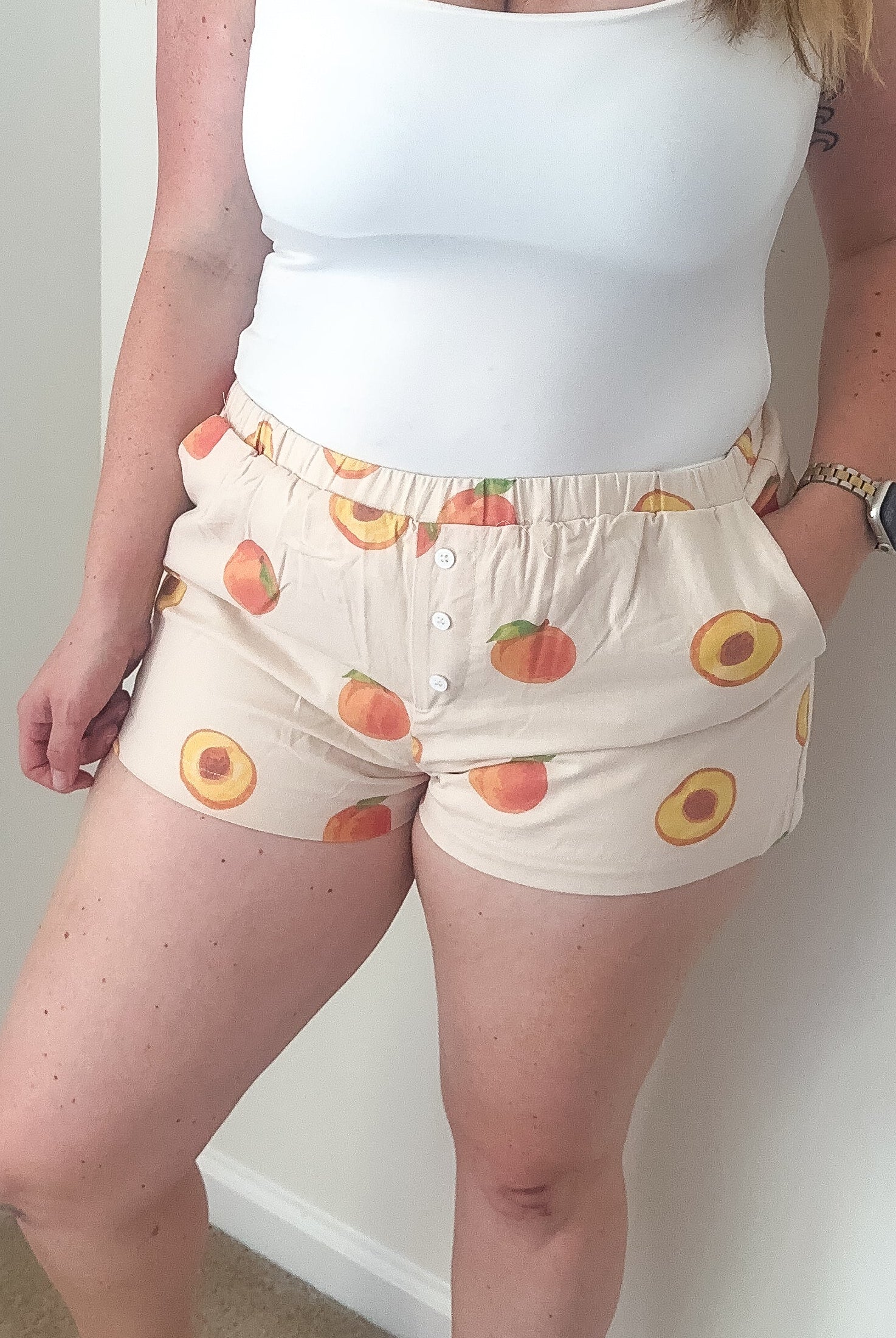 Woman wearing a white tank top and patterned shorts against a plain wall.