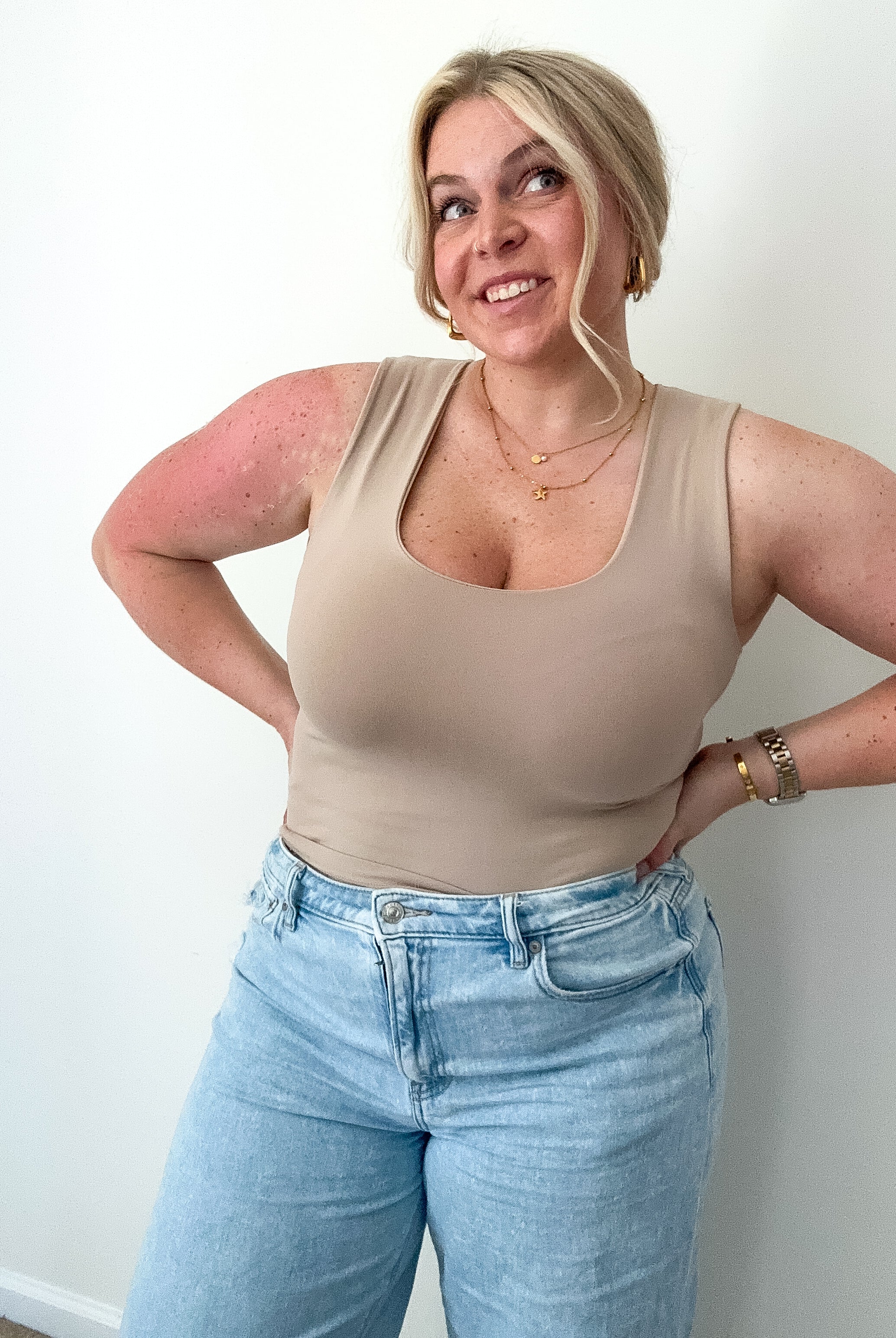 Woman wearing a beige tank top and light blue jeans against a white wall