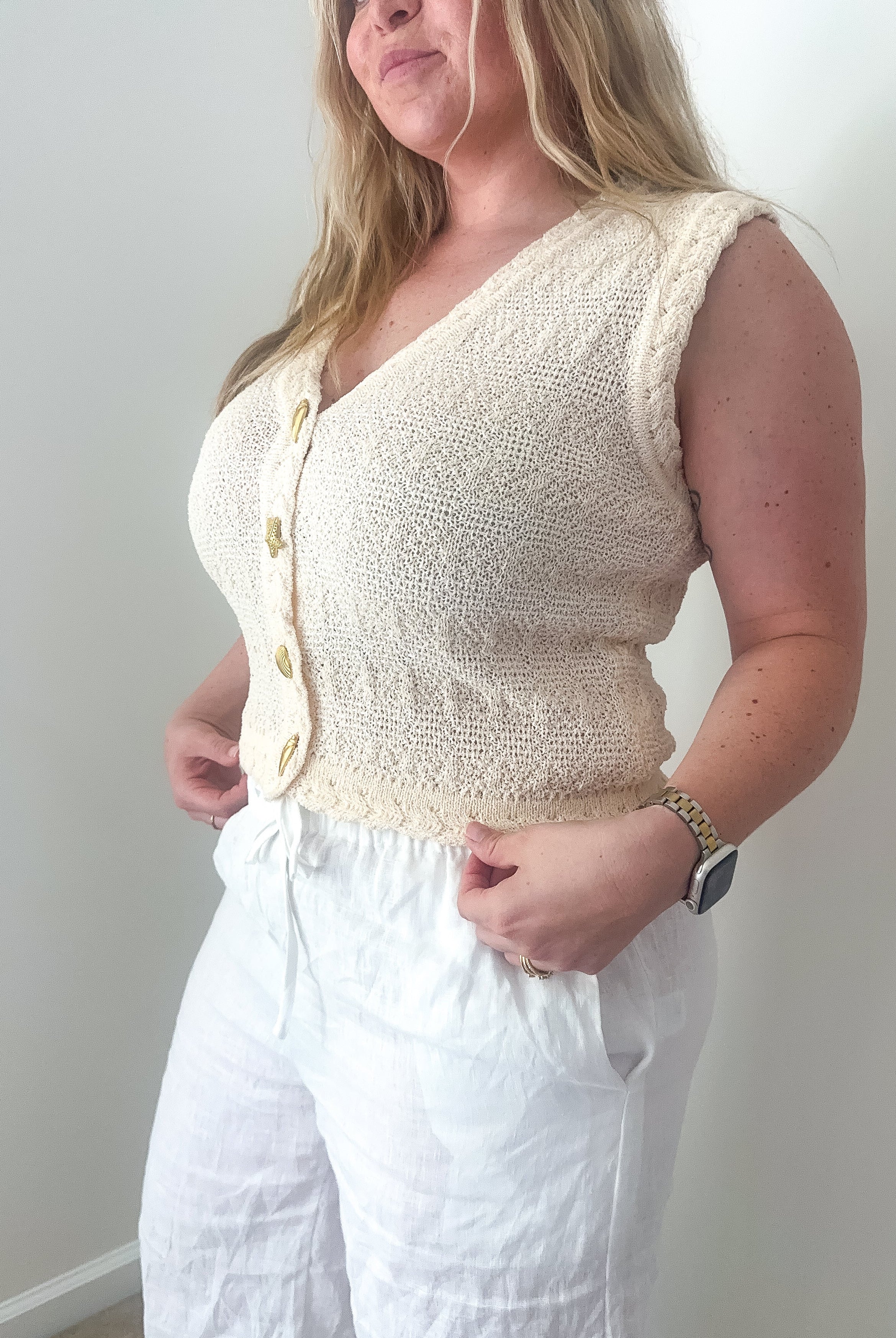 Woman wearing a cream-colored sleeveless knitted top and white pants against a plain background
