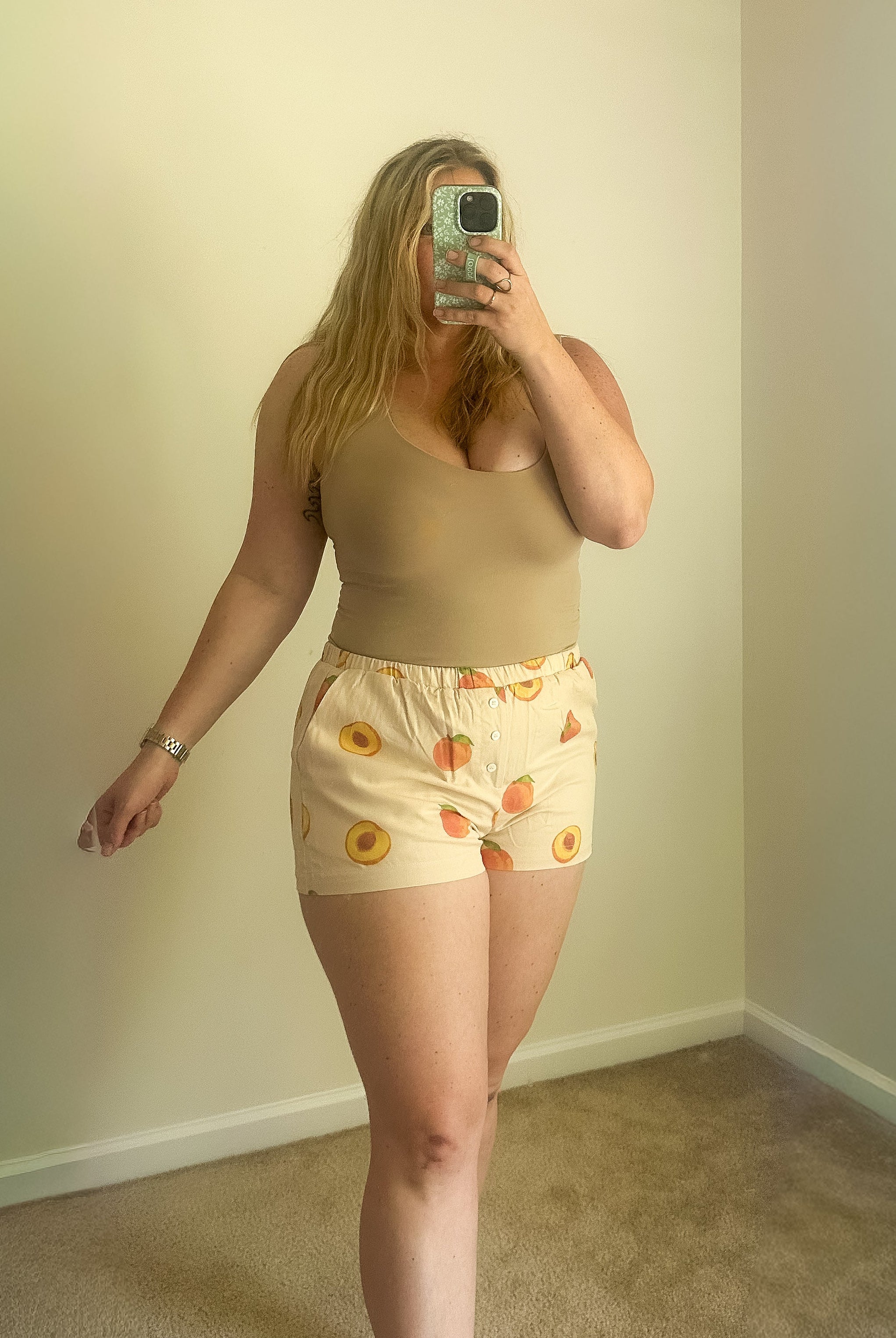 Person taking a mirror selfie wearing a beige top and patterned shorts in a room with plain walls.