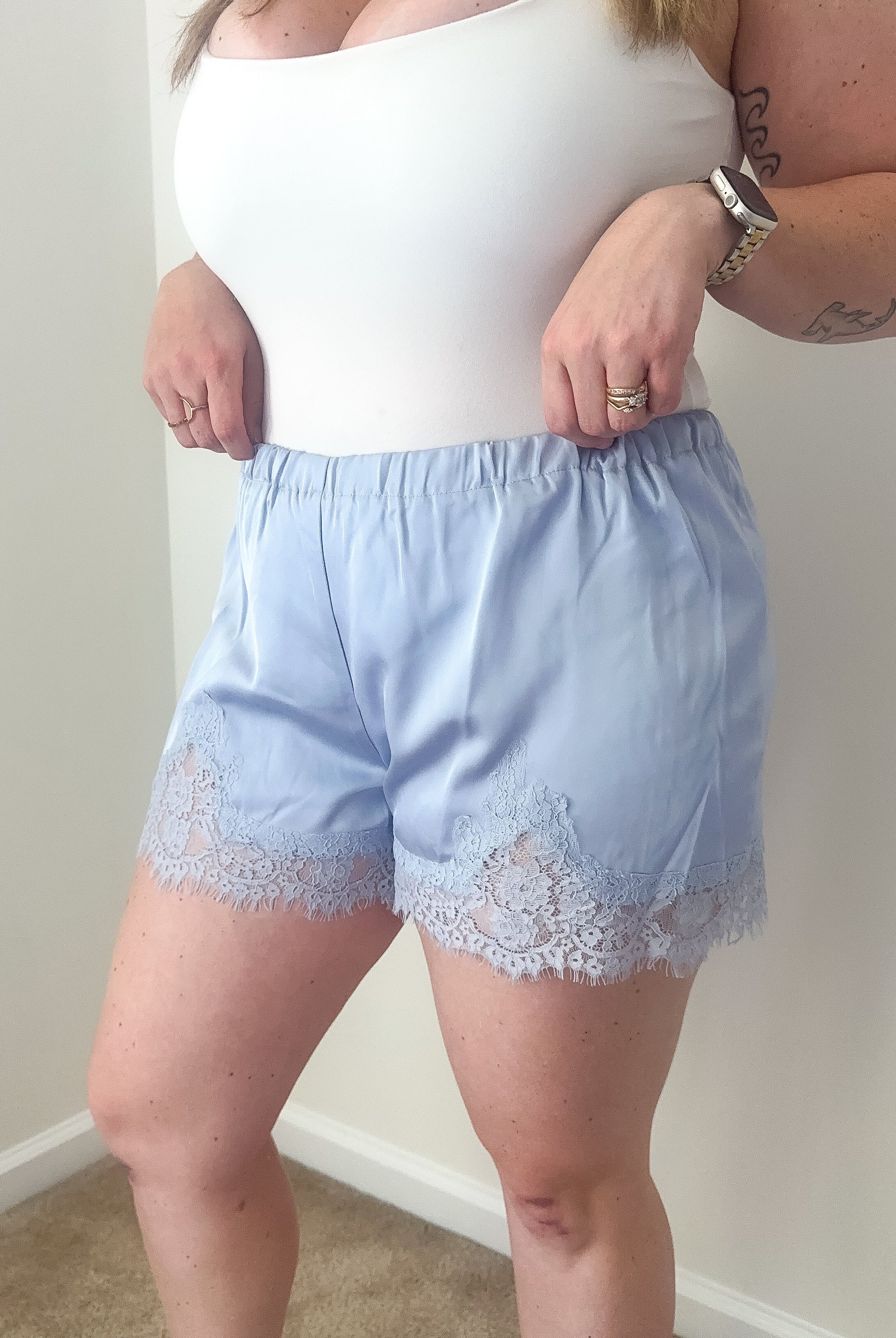 Person wearing a white top and light blue shorts with lace trim against a plain wall.