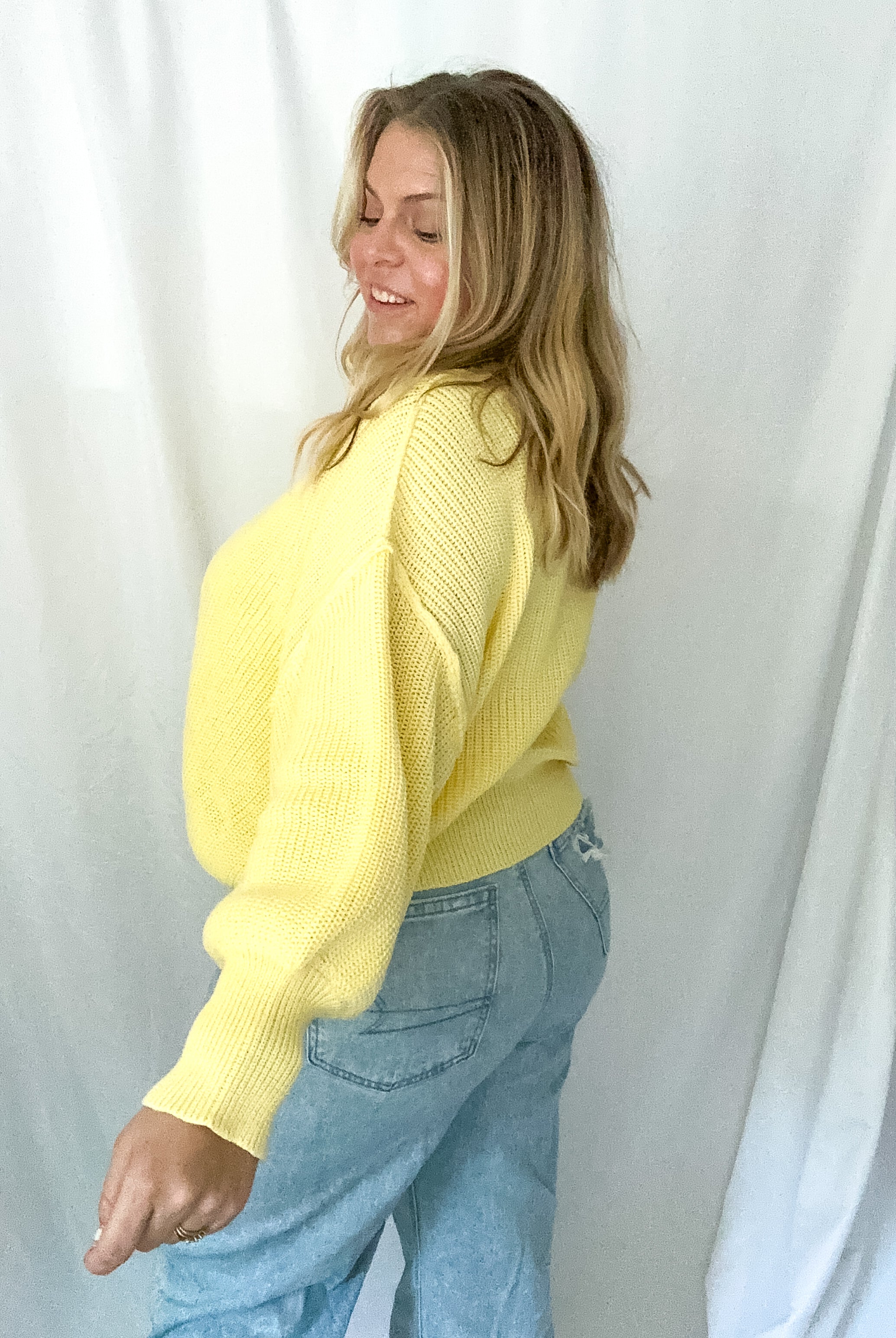 Woman wearing a yellow sweater and blue jeans against a white curtain background