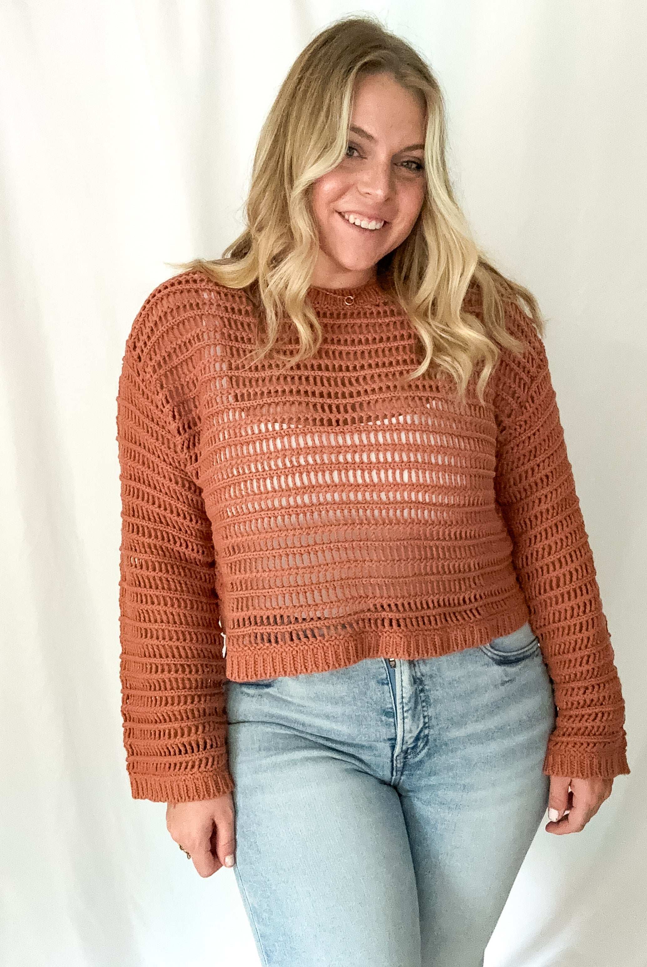 A woman wearing a rust-colored openwork crochet sweater with long sleeves and a boat neckline.