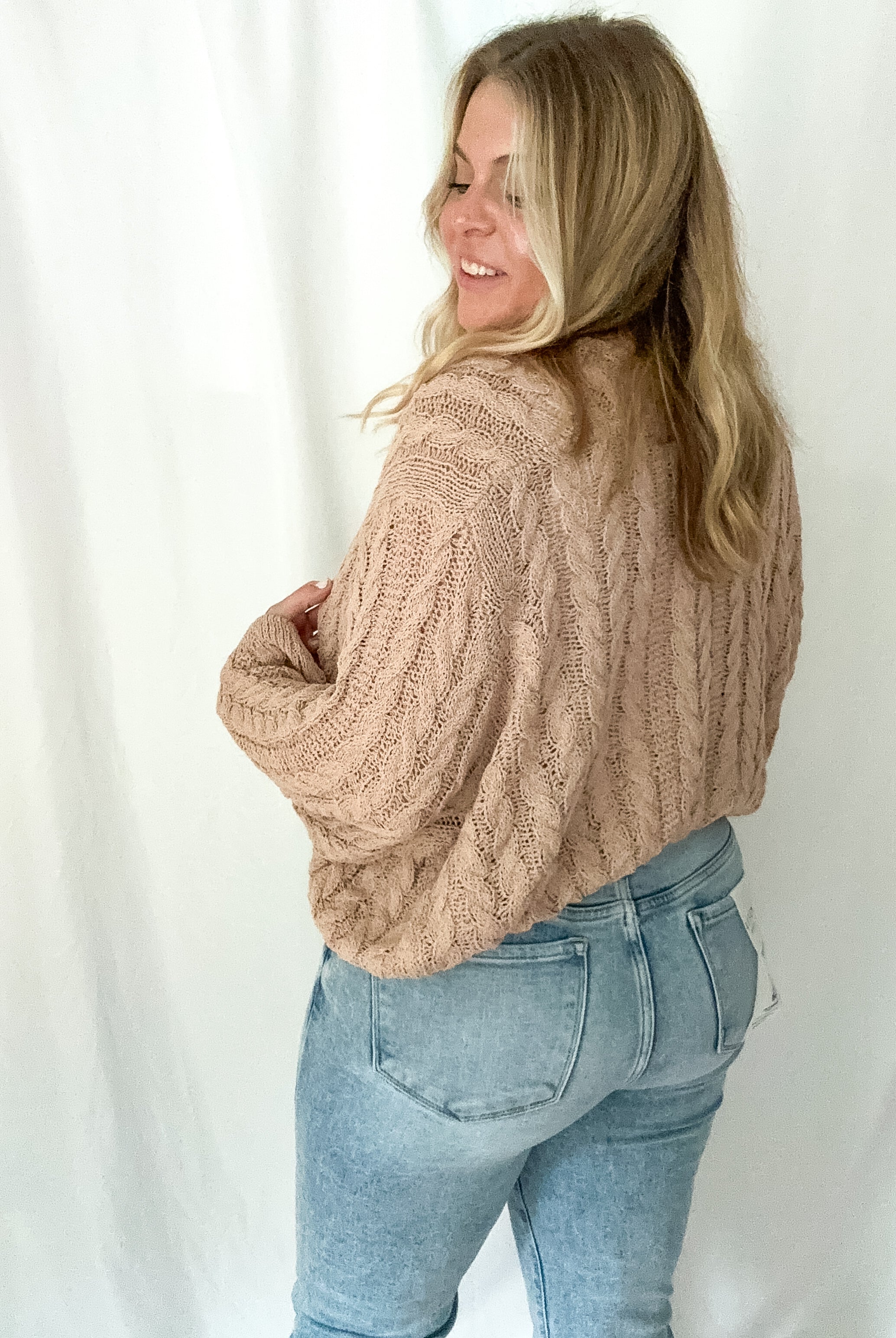 Woman wearing a beige cable knit sweater and light blue jeans against a white background