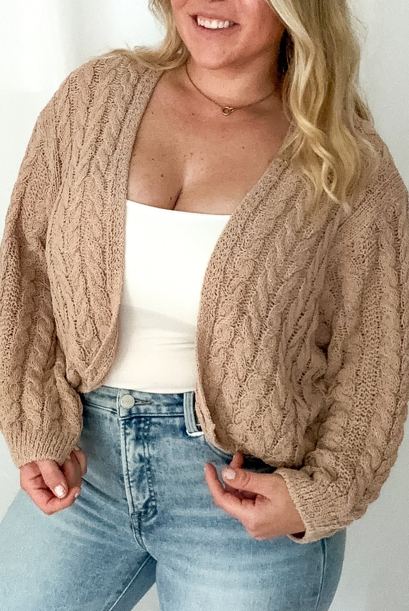 Woman wearing a beige cable knit cardigan over a white top and light blue jeans on a plain background