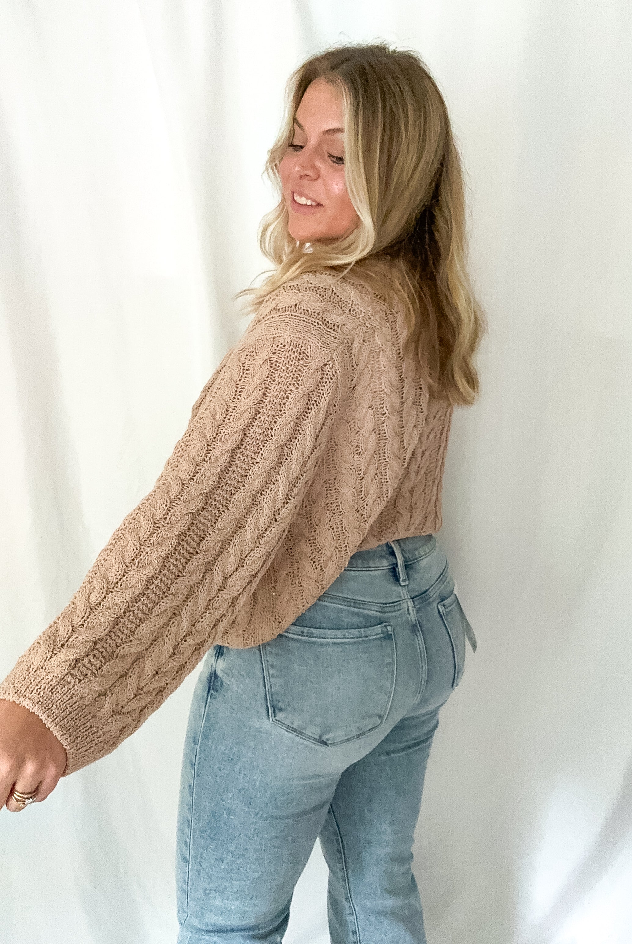 Woman wearing a beige cable knit sweater and blue jeans against a white background