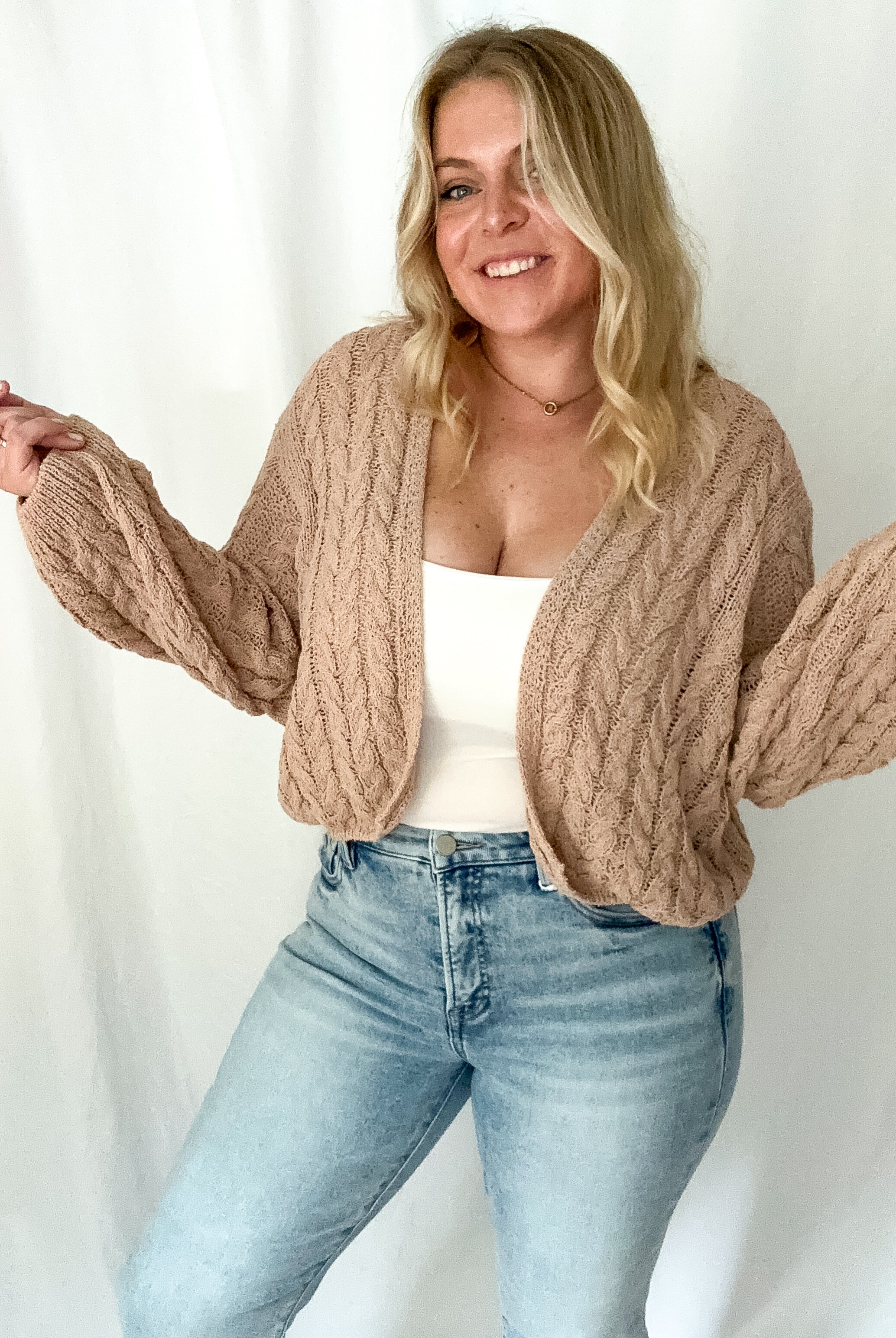 Woman wearing a beige cable knit cardigan over a white top and blue jeans on a white background