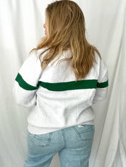 Person wearing a white sweater with green stripes and light blue jeans against a white curtain background