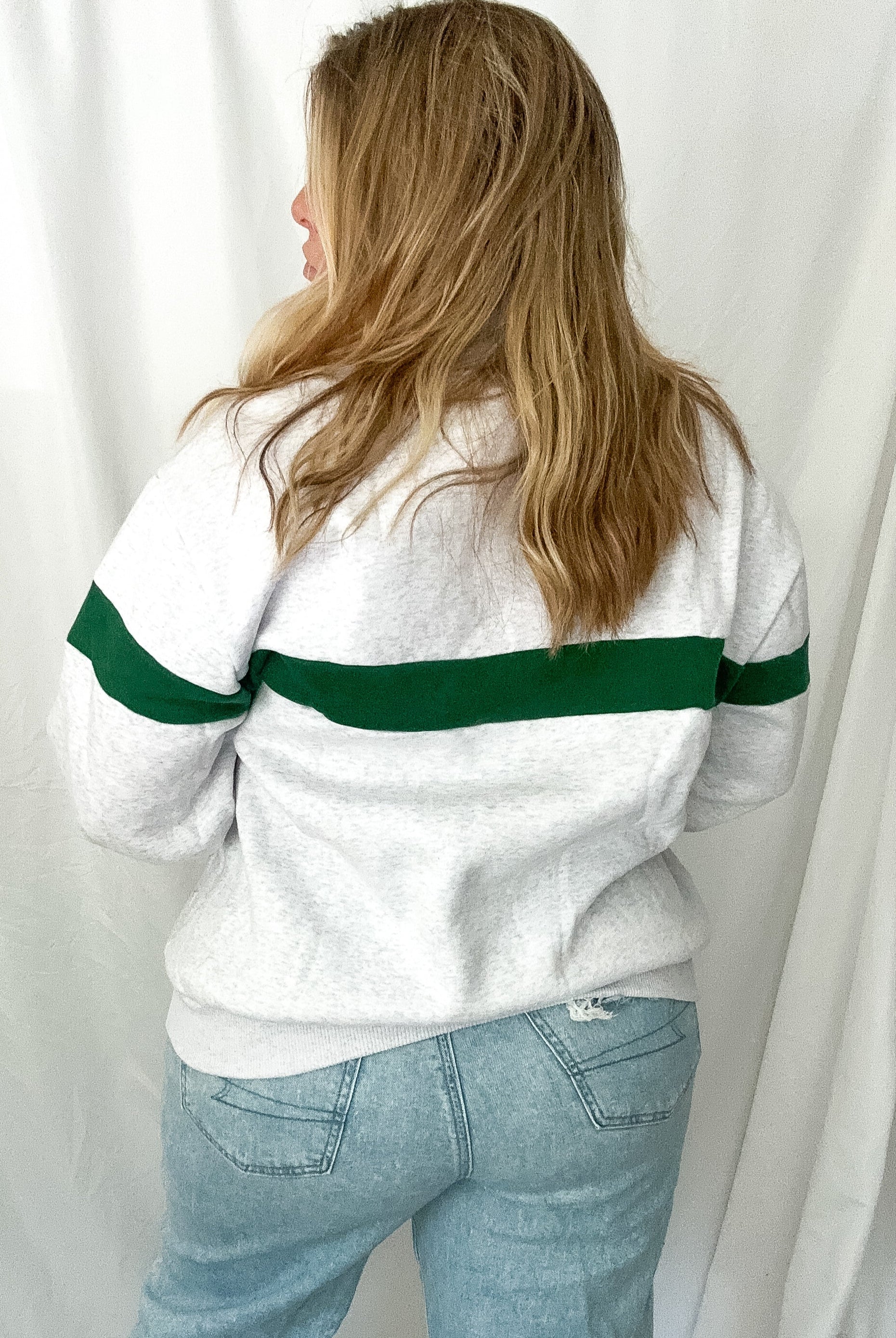 Person wearing a white sweater with green stripes and light blue jeans against a white curtain background