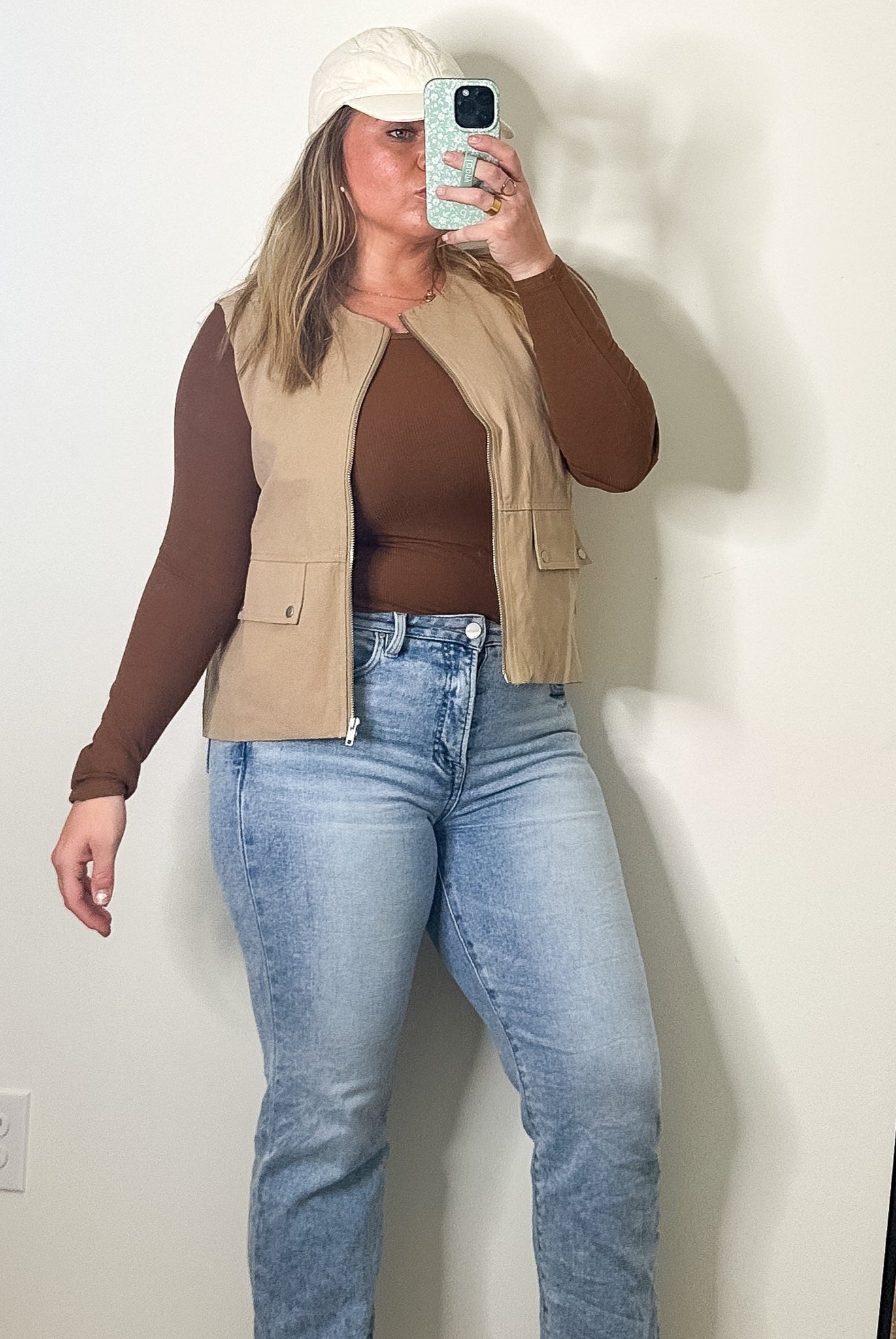 Person taking a mirror selfie wearing a brown top, beige vest, and blue jeans in a room with a large mirror.