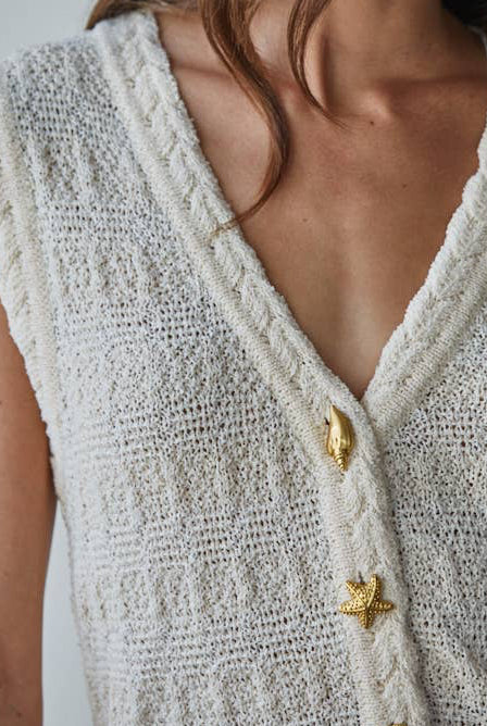 Woman wearing a textured white sleeveless top with gold buttons against a plain background
