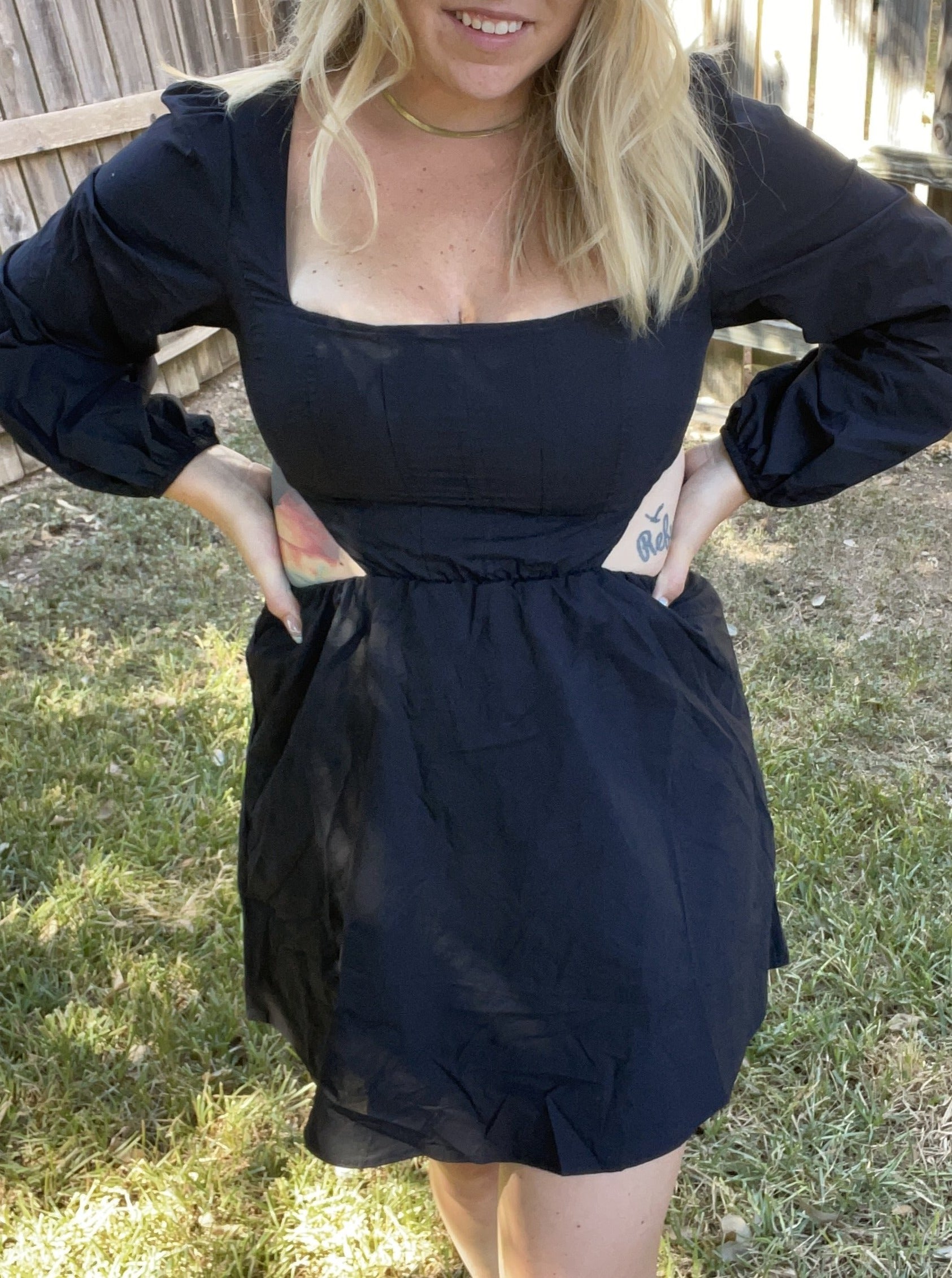 A woman wearing a black, short-sleeved, V-neck dress with a cut-out detail on the back, standing in an outdoor setting.