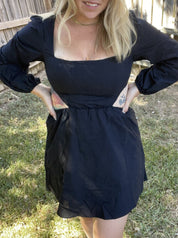 A woman wearing a black, short-sleeved, V-neck dress with a cut-out detail on the back, standing in an outdoor setting.