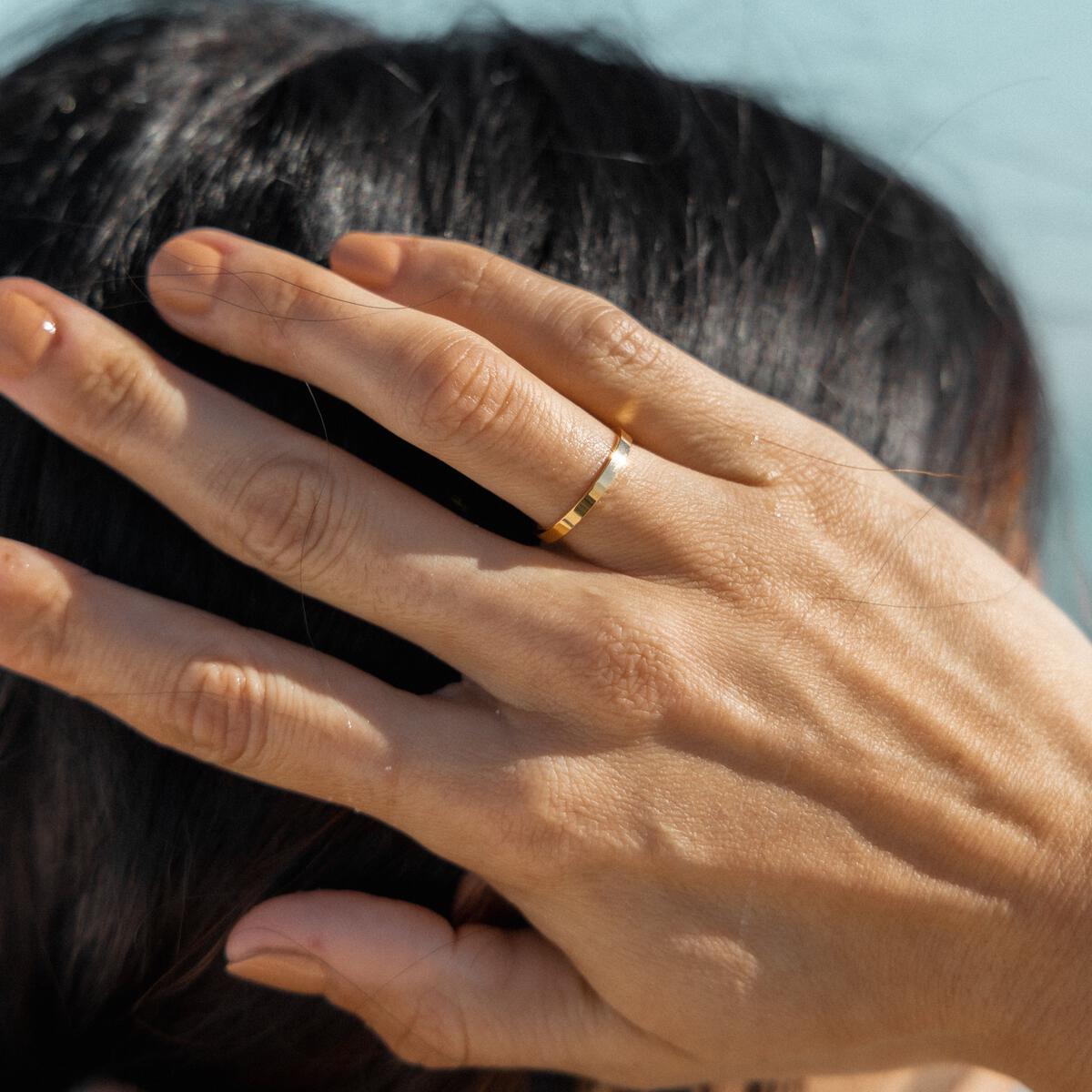 A hand wearing a simple gold-plated ring against a coastal backdrop.
