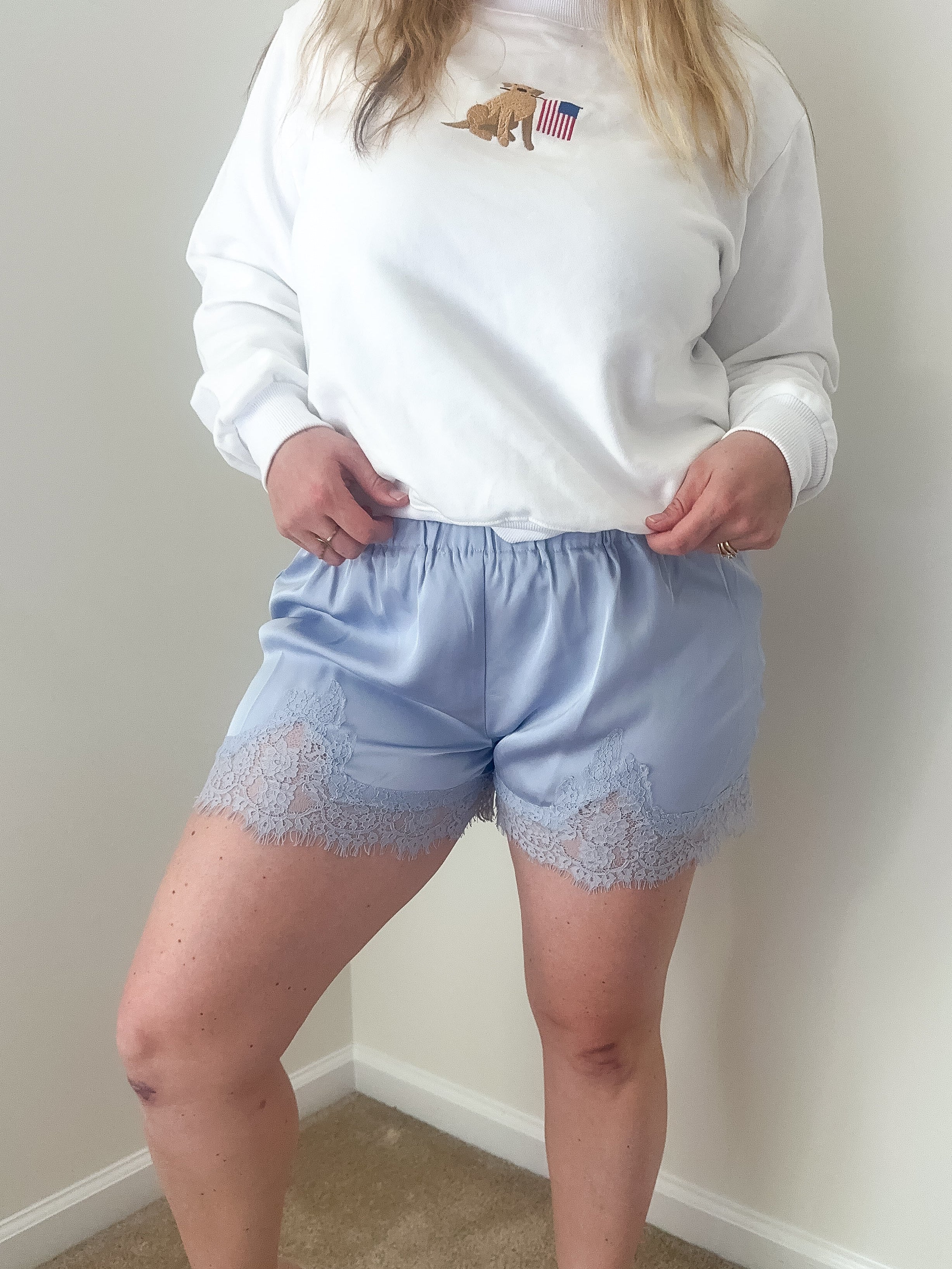 Person wearing a white sweatshirt with a logo and light blue shorts with lace trim against a plain wall.