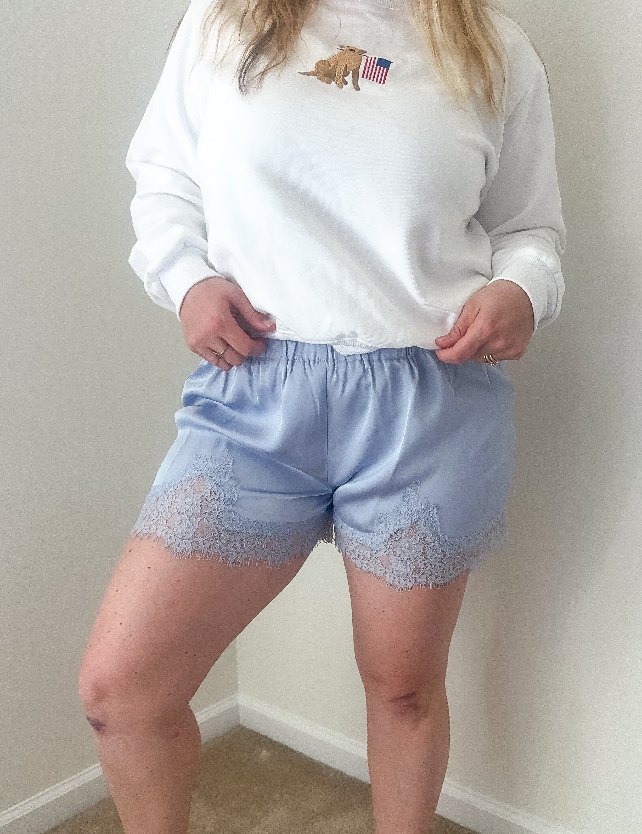 Person wearing a white sweatshirt with a logo and light blue shorts with lace trim against a plain wall.