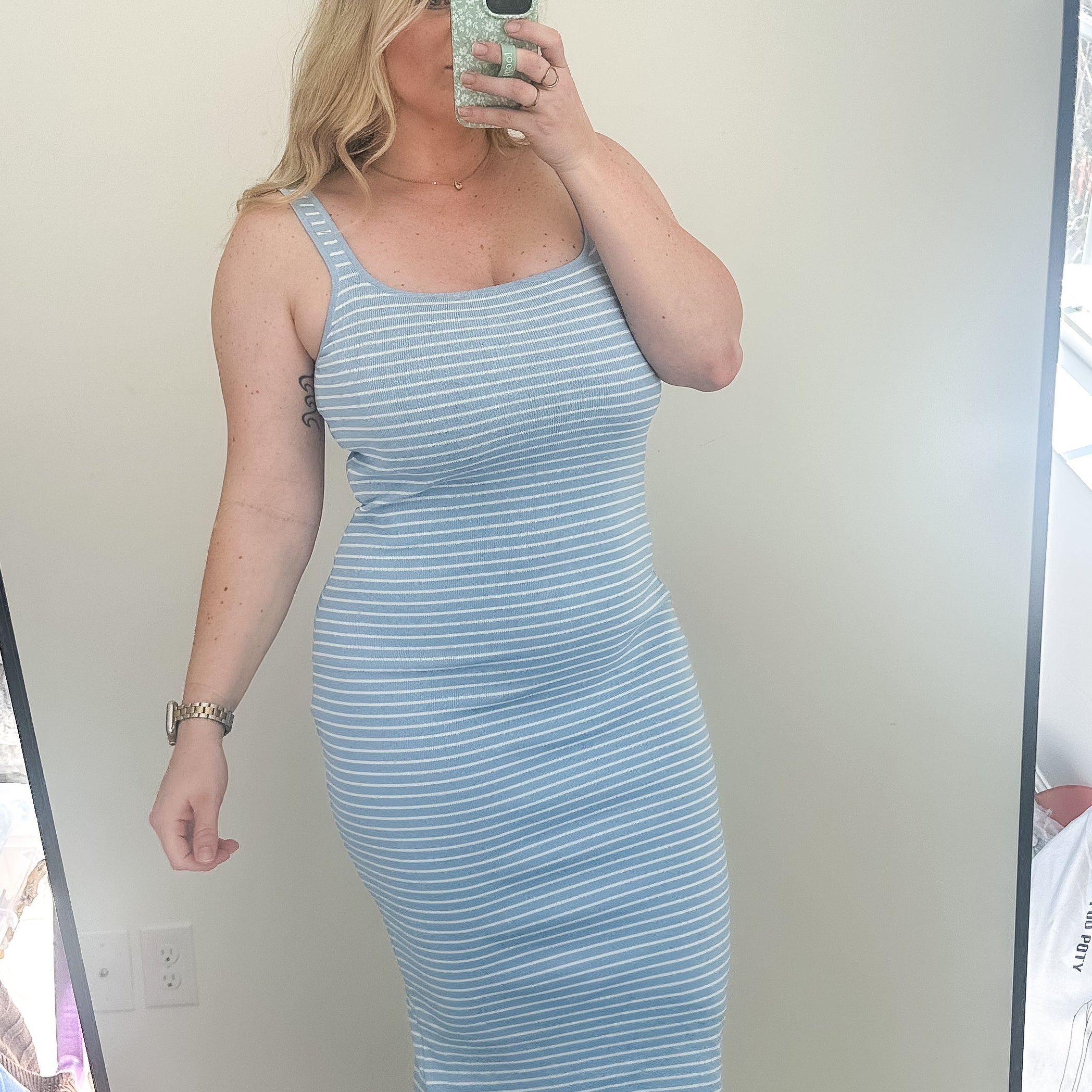 Woman taking a mirror selfie wearing a light blue striped dress in a room with a window.